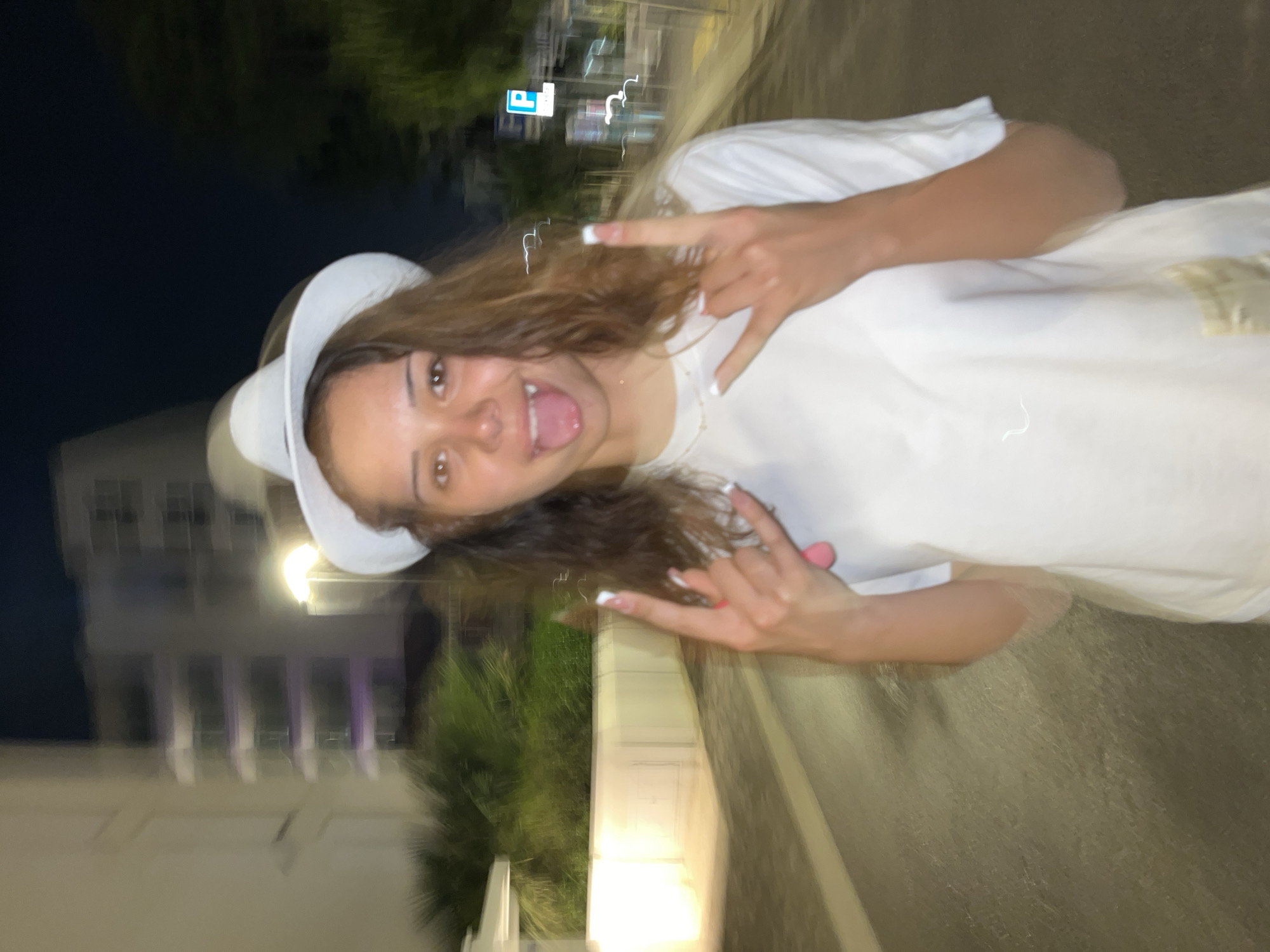 A woman with medium-length brown curly hair, wearing a white T-shirt and a white hat, is making the rock-on hand gesture with both hands. Her tongue is sticking out, and she is looking at the camera. The background shows an outdoor scene in what appears to be Jesolo, Italy at night, with blurry buildings, trees, and streetlights visible.