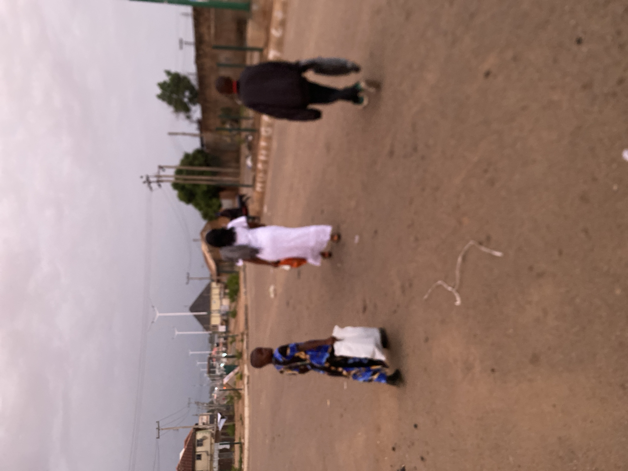 Three individuals are walking along a paved road in Bukuru, Nigeria. One person wears a dark jacket. A second person is in white clothing. The third individual wears a blue and yellow patterned outfit. Streetlights and buildings are visible in the background. Overhead power lines are also present.