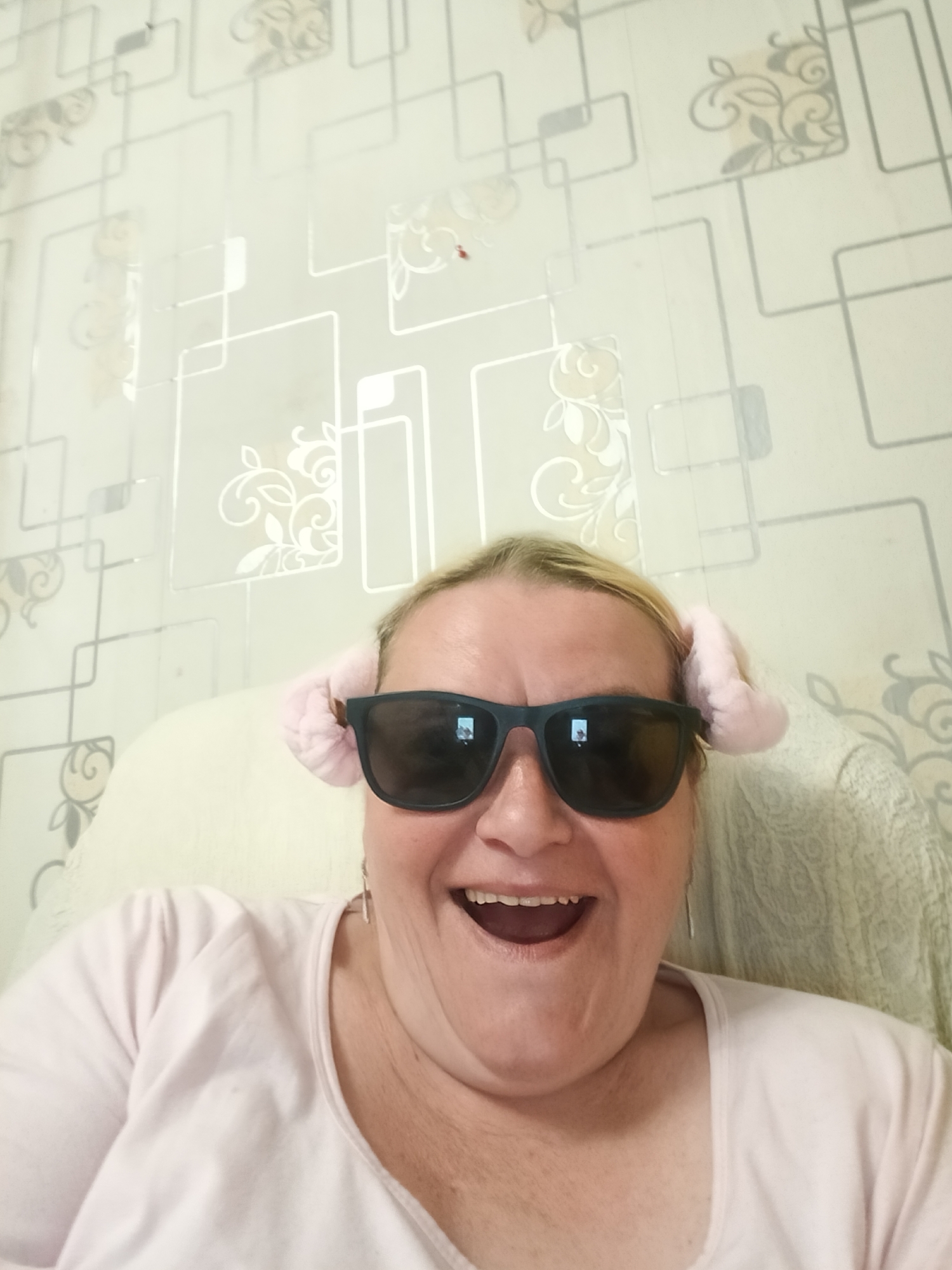 A woman with blonde hair, wearing dark sunglasses and pink bunny ear headbands, is smiling widely. She is wearing a light pink shirt. The background shows a patterned wallpaper with a design of squares and floral motifs. She appears to be sitting in a chair or couch. The location is Красный Маяк, Russia.