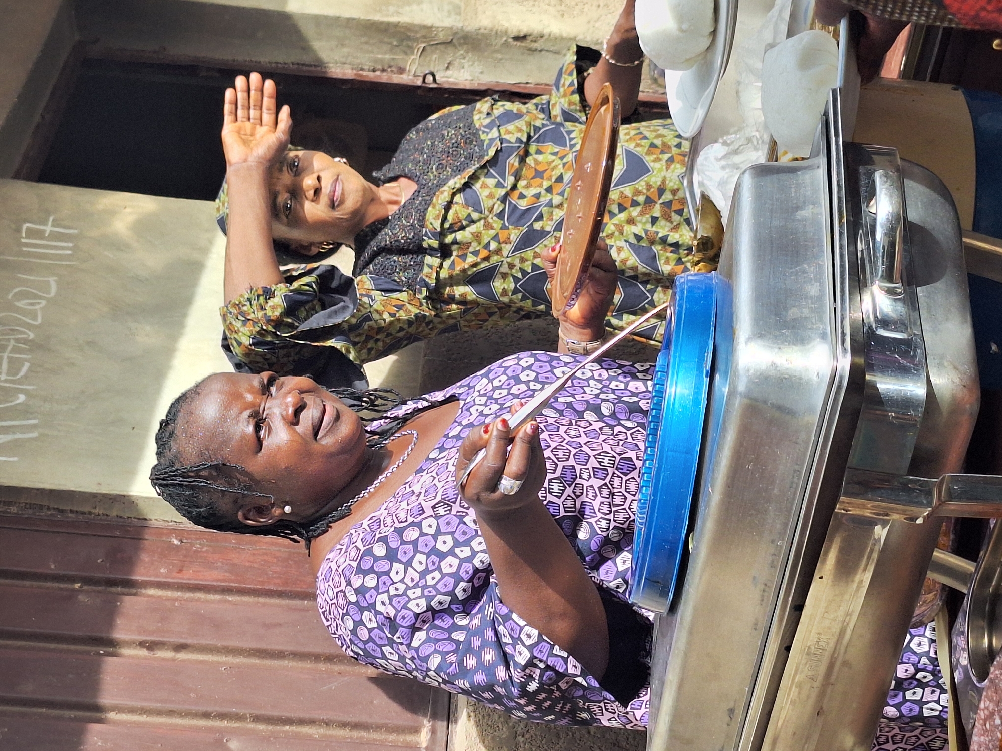 Two African women stand in Sabon Tasha, Nigeria, near a metal counter with white, dough-like objects on plates and a blue-lidded container. One woman wears a purple patterned dress and holds a metal utensil. The other woman wears a patterned dress, holds a plate, and raises her left hand. Writing is on a light-colored wall nearby.