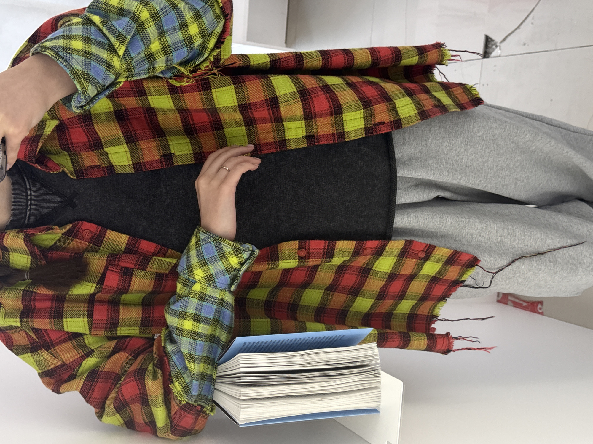 The image shows a person sitting at a white table reading a book. The person is wearing gray sweatpants, a gray top, and a brightly colored plaid shirt in shades of yellow, red, and blue. Their hands are resting on the book, and a wedding ring is visible on their left ring finger. The book has a light blue cover and many white pages. The background is a white wall with a small white object on it.