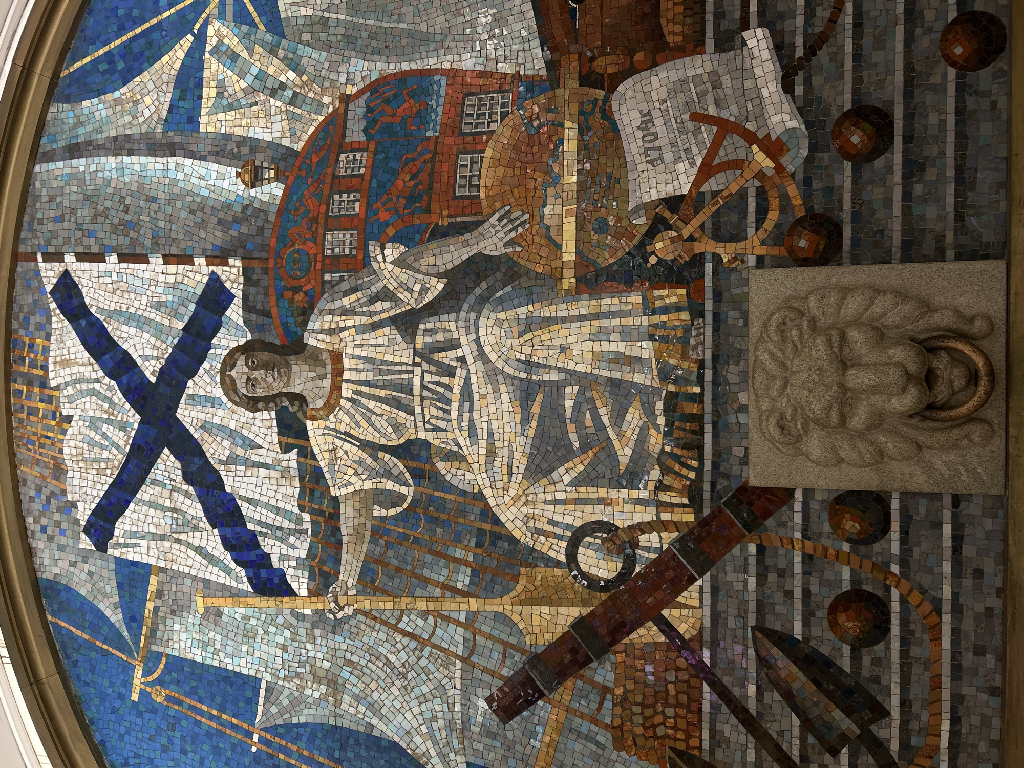 This is a mosaic mural found in the St. Petersburg Metro in Russia. The scene depicts the founding of St. Petersburg by Peter the Great. Peter the Great stands centrally, surrounded by imagery representing the founding of the city, including a building, a naval flag, navigational tools, and architectural plans. A stone lion head adorns the right of the mosaic.