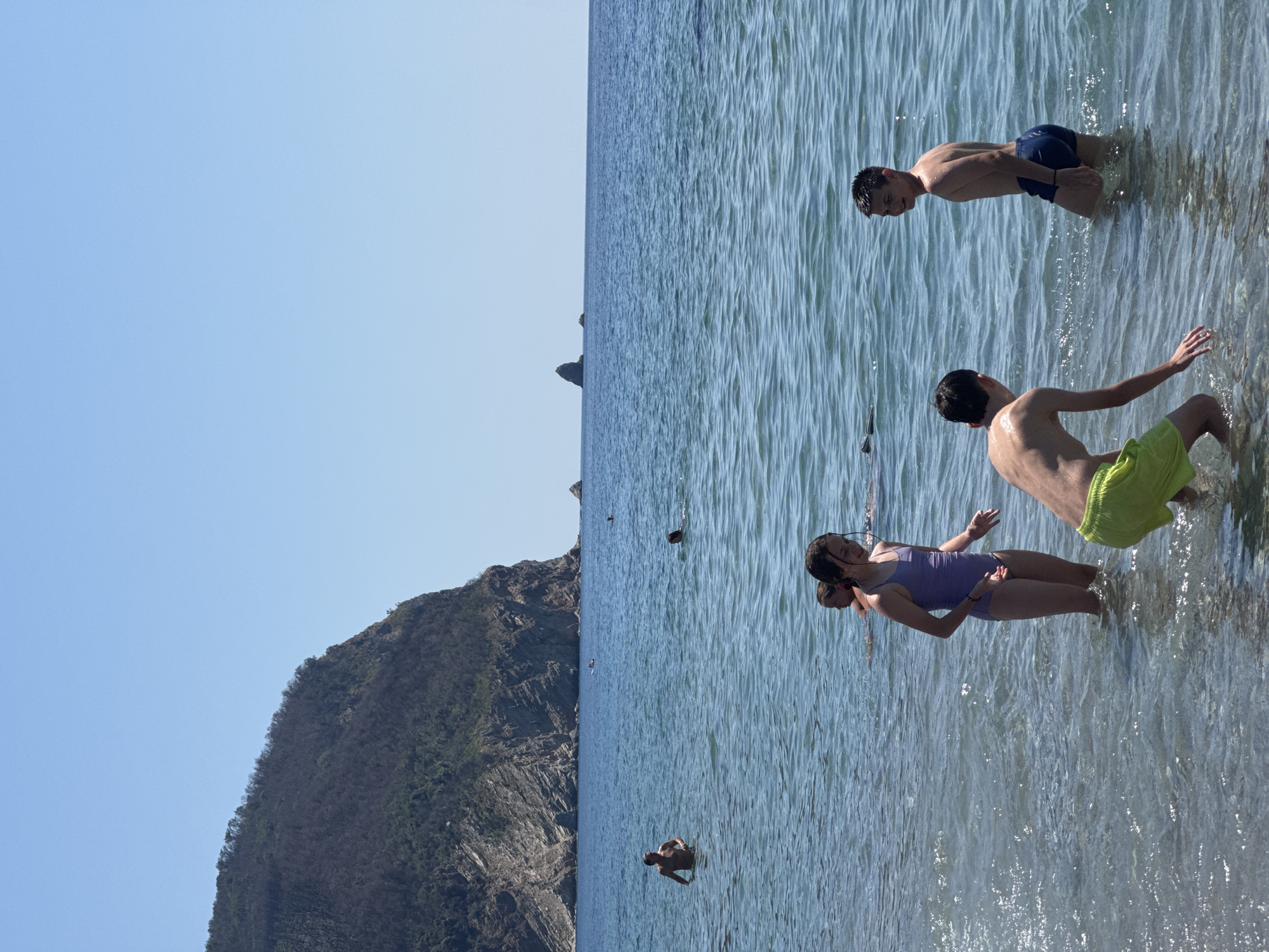 This is a full-shot, vertical, natural-light photograph taken from the water's edge, looking out towards the sea. In the foreground, three children are in the water. On the left, a boy in bright green swim trunks bends down, facing away from the camera. To his right, a girl in a purple swimsuit stands, looking to her left with her hands slightly outstretched. To the far right, a boy in dark blue swim trunks is partially submerged, looking over his right shoulder towards the camera. In the middle ground, several other people are visible in the water, swimming or wading. In the background, a large, rocky, tree-covered mountain juts out into the sea on the left side of the frame. The sea is a clear blue with gentle ripples. The sky is a bright, cloudless blue. The scene appears to be the coast of Đurovići, Montenegro.
