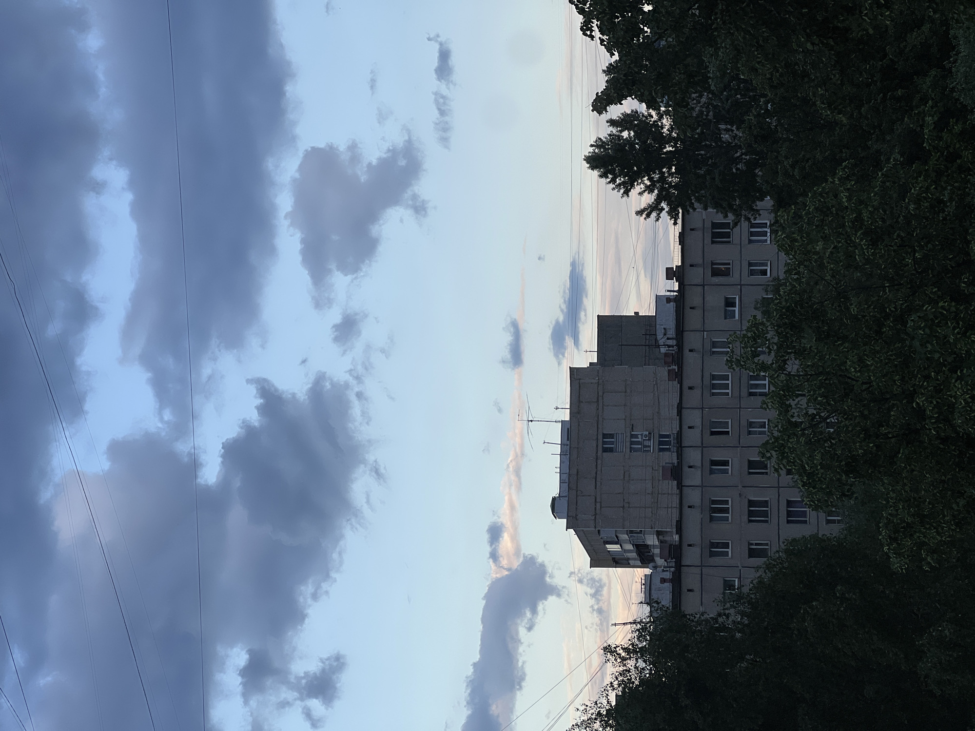 A photograph depicts an overcast sky above Khimki, Russia, and a Soviet-era apartment building. The clouds are mostly grey and dark, suggesting potential rain. Power lines run diagonally across the sky in the upper left. The apartment building is constructed of concrete. A dense canopy of trees partially obscures the building's facade. There are no people visible.