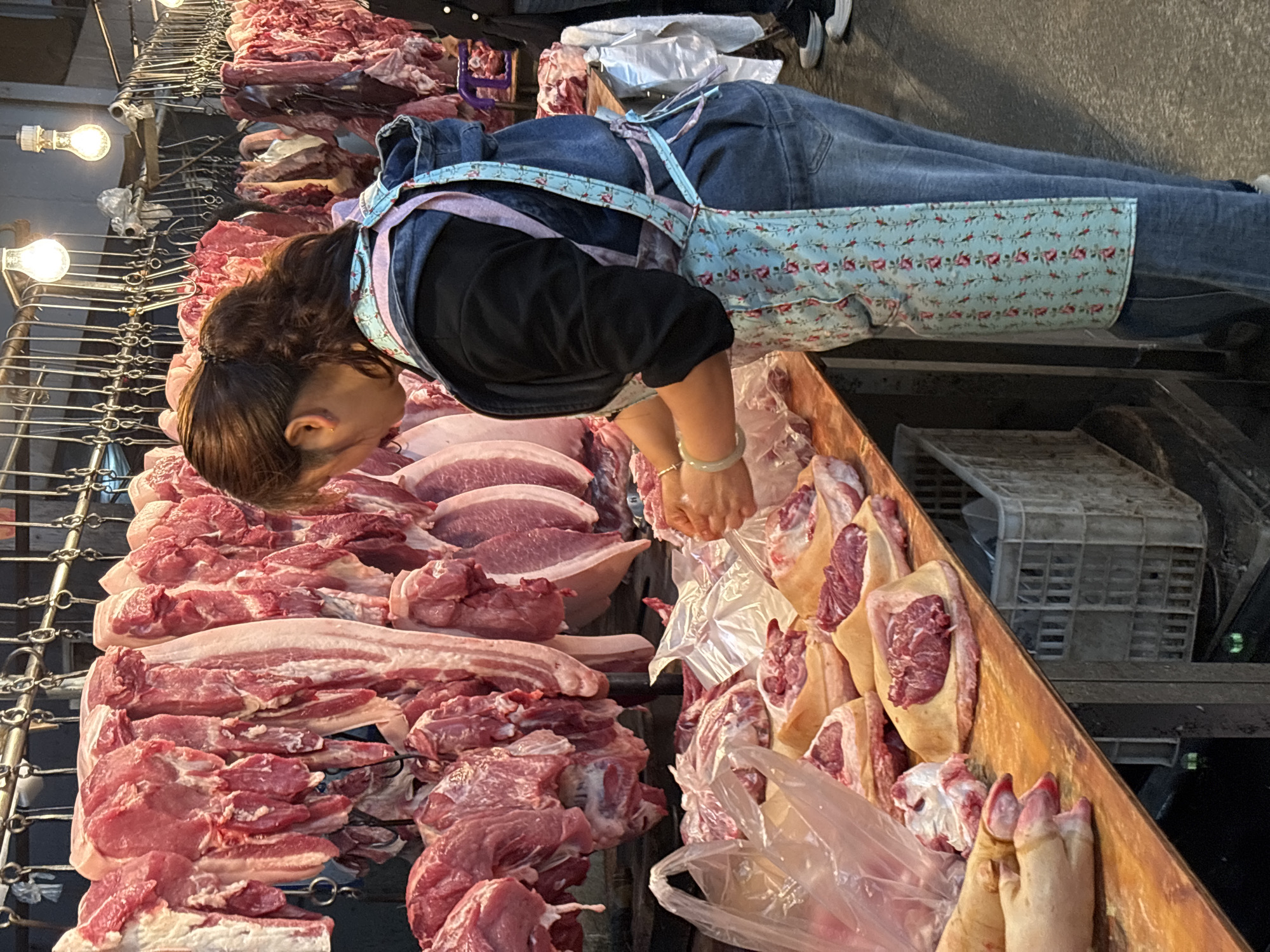A woman wearing jeans, a black top, and an apron with a floral pattern, stands behind a table laden with raw meat, likely at a meat market. She is sorting the meat, some of which is packaged in plastic bags. Chains with meat hanging from them are above the table. A plastic crate is visible behind the table. The presence of the meat market suggests this scene is potentially located in Jiangjin, China, due to the prevalence of these types of markets throughout China.