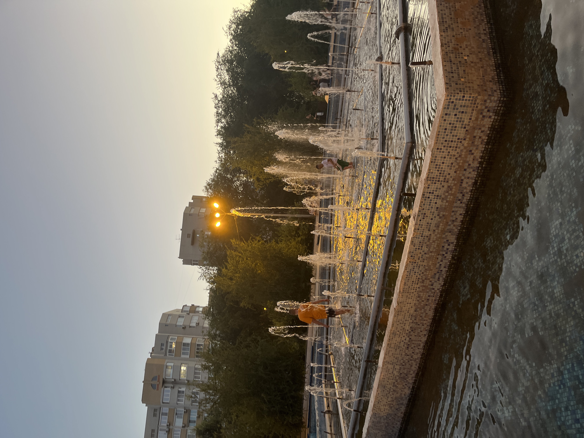 The image depicts a public fountain area in Astrakhan, Russia, during twilight. Two young boys are visible in the fountain, one in an orange shirt and dark shorts, the other in a white shirt and green shorts. They are wading through the water, surrounded by numerous jets of water spraying upwards. In the background, trees line the edge of the fountain area, with multi-story apartment buildings visible beyond the trees. Streetlights illuminate the scene with a warm glow. The sky is clear and transitioning from daylight to evening. The fountain itself has tiled edges and a complex arrangement of water jets. Several other individuals are indistinctly visible in the background near the trees, suggesting a public gathering space.