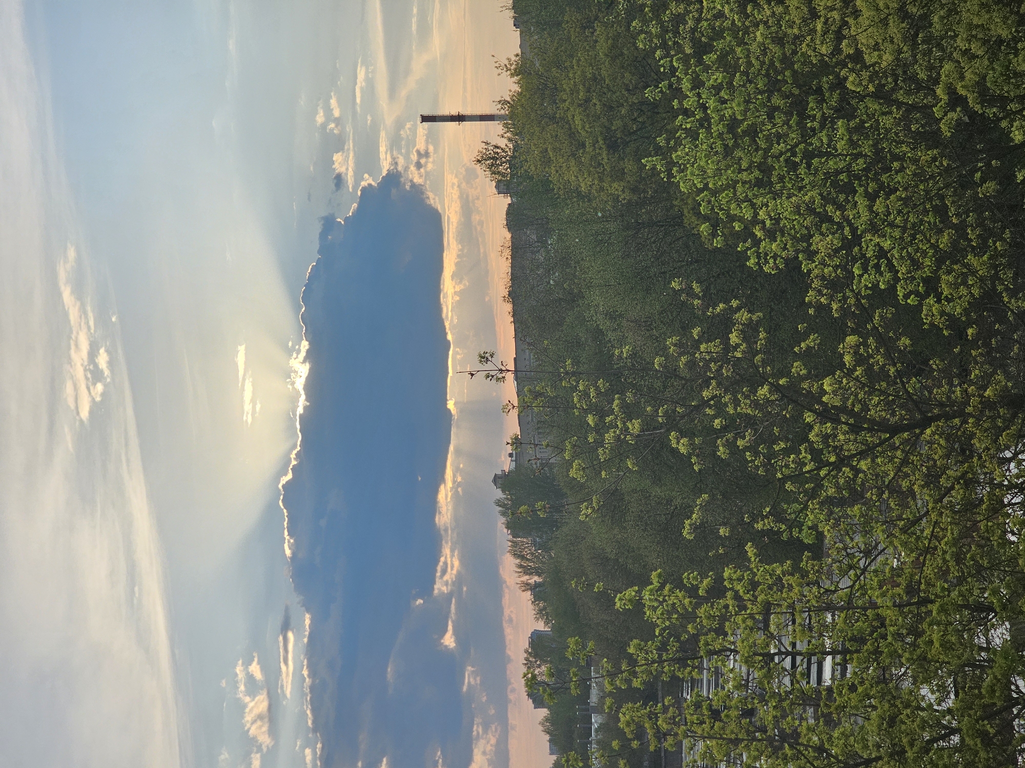 The image shows a scene in Moscow, Russia, likely captured during sunset or sunrise. In the sky, a large, dark cloud partially obscures the sun, creating rays of light that break through its edges. Other clouds are scattered around the sky, with hues of blue and faint orange indicating the time of day. Along the horizon, there is a line of green foliage, presumably trees, interspersed with buildings. A tall smokestack is visible in the distance. The foreground shows more greenery from trees, partially obscuring the buildings below.