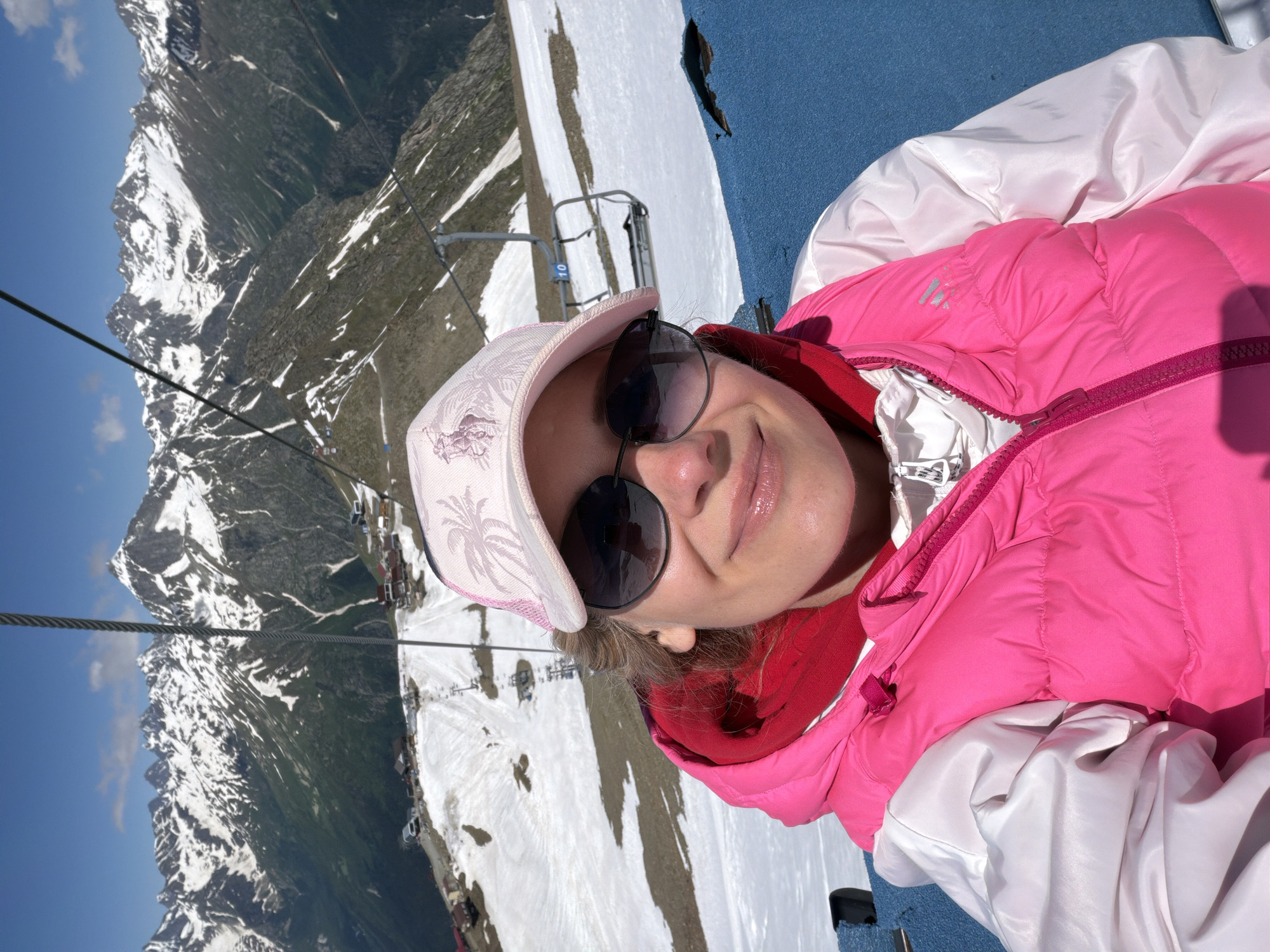 A medium shot selfie shows a woman wearing a pink puffer vest over a white jacket and a pink baseball cap, looking at the camera. She is wearing sunglasses. The background is a mountain landscape with snow-capped peaks and a ski lift. The location is a ski resort in Верхне-Тебердинское сельское поселение, Russia. The sky is clear blue with some scattered clouds. The overall scene is bright and sunny.