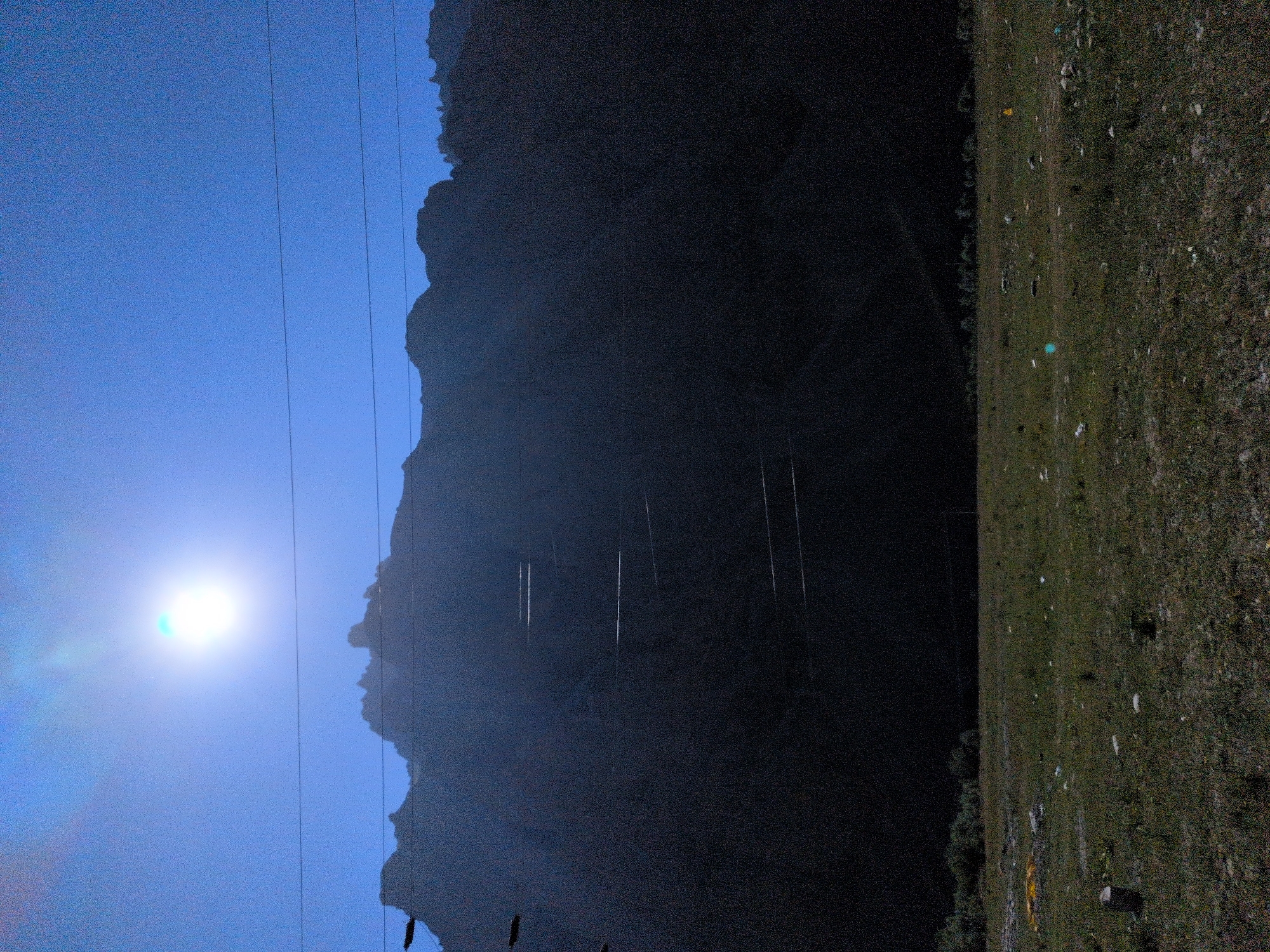 The image depicts a vast, dark mountain range silhouetted against a bright, hazy sky. The sun is a prominent, almost blinding white orb in the upper left quadrant, casting a strong glare. Several thin, dark power lines stretch horizontally across the frame, partially obscuring the mountain's features. These lines are strung between unseen poles, creating a sense of infrastructure within the natural landscape.
The mountain itself is largely devoid of detail due to the low light and shadow, appearing as a solid, dark mass. However, a series of lighter, horizontal stripes are faintly visible on its face, suggesting man-made terracing or possibly reflective elements catching the light. The terrain at the base of the mountains transitions into a grassy field, dotted with scattered rocks and debris. The grass is a mix of green and brown, indicating a dry or somewhat sparse growth. In the distance, beyond the field, a line of trees or shrubs is visible, forming a dark, irregular border. The overall atmosphere is one of a stark, natural landscape with subtle human intervention. The location is identified as Matayeen, India.