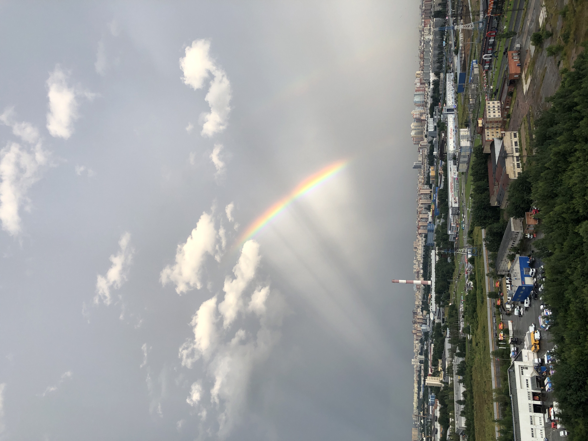 A panoramic view showcases a city under a dramatic sky. A prominent rainbow arches across the frame, its vibrant colors a stark contrast to the overcast, gray clouds that dominate the upper portion of the image. Sunbeams pierce through the clouds, creating visible shafts of light. Below the sky, a sprawling urban landscape unfolds. A tall, industrial smokestack with red and white bands stands out against the horizon, a symbol of industry. The city itself is a dense collection of buildings, ranging from low-rise industrial structures to taller residential buildings in the distance. Green trees and foliage are interspersed throughout the urban sprawl, particularly in the foreground where a dense line of trees partially obscures a cluster of industrial buildings and parked vehicles. The scene appears to be captured during daylight hours, likely after a rain shower, given the presence of the rainbow and the subdued lighting. The overall impression is one of an industrial city experiencing a moment of natural beauty.