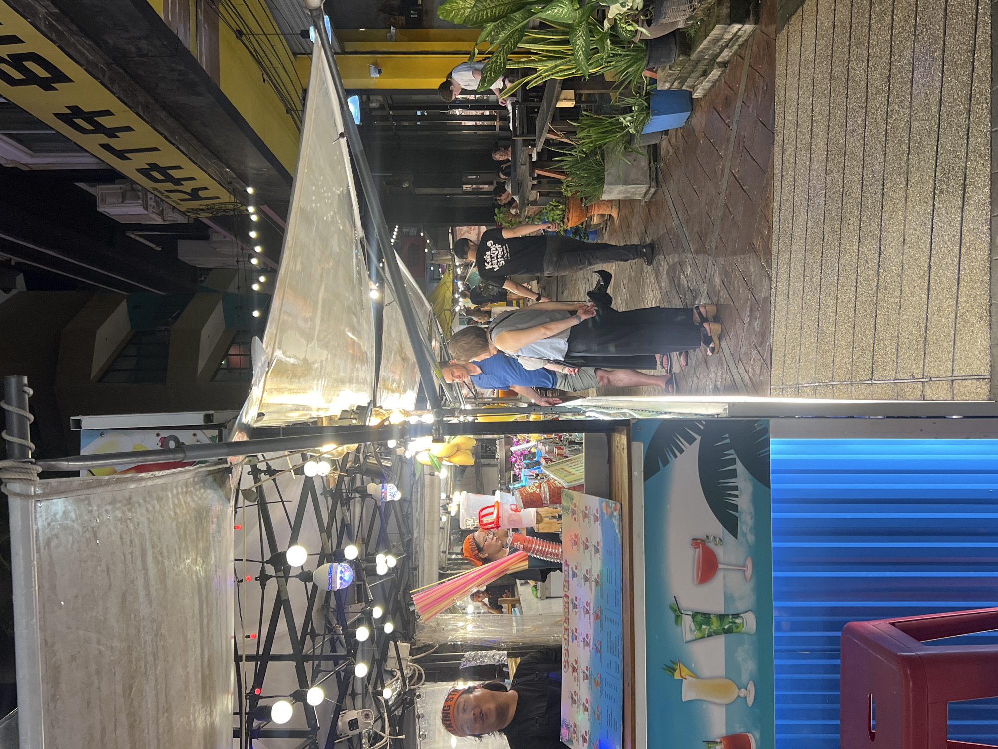 The image captures a vibrant street scene in Karon, Thailand, likely in the evening. A cocktail bar stall is prominently featured, displaying a colorful menu with illustrations of drinks. Two individuals, possibly employees, are visible behind the counter, one wearing a headband and the other an orange bandana. Several customers are seen walking along the pavement and seated at tables in the background. The street is lined with buildings, one of which has a large yellow sign with black lettering that reads "KATA BUNG." Potted plants and string lights enhance the atmosphere, suggesting a lively commercial area. The ground appears wet, possibly from recent rain.