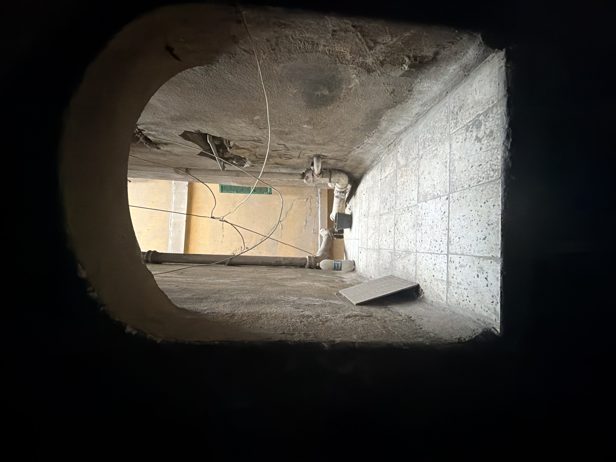 The image captures a view through an arched opening into a narrow, outdoor alleyway or courtyard in Cairo, Egypt. The scene is dimly lit, with the foreground heavily shadowed by the archway.
The alleyway is paved with square, light-colored tiles that show some wear. The walls on either side are rough and textured, appearing to be made of aged stucco or concrete. The right wall has a visible patch of damage and some dark staining. A vertical pipe runs down the left side of the alley, connected to a horizontal pipe that runs along the ground.
Various objects are present: a white plastic bottle with Arabic writing on its label stands near the pipe on the right side. To its left, a square, textured metal plate leans against the wall. Further down the alley, a black bucket is visible near the pipes, and beyond that, a green shuttered window can be seen on a pale yellow building in the distance. Several thin wires are strung across the alleyway and attached to the walls. The overall impression is of a utilitarian, somewhat neglected urban space.