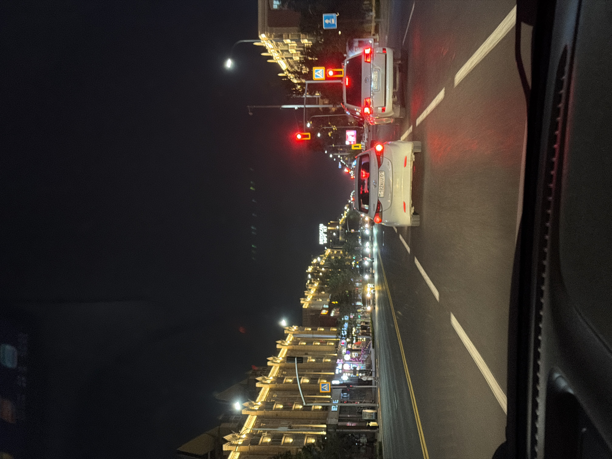 A nighttime view from a vehicle looking out onto a city street in Astana, Kazakhstan. The street is illuminated by streetlights and the glowing lights of buildings lining the left side of the road. A row of light-colored buildings with ornate architecture and yellow lighting stands prominently. Traffic lights are visible, with red lights illuminated for vehicles. Several cars are on the road, including a white sedan in the foreground and a white SUV to its right. The road has clear lane markings. The dark night sky is visible above, with some faint green light streaks.