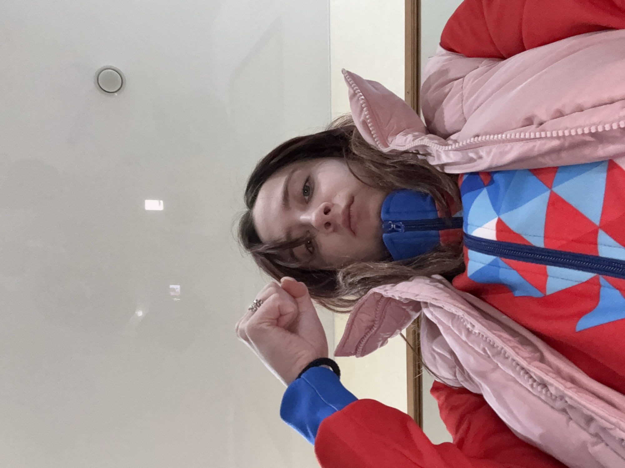 The image shows a woman with brown hair wearing a red, blue, and light pink winter jacket. She is holding up her left fist, where she wears a ring. She is indoors with a white ceiling. Based on the information, it is not possible to ascertain her location in Gory, Russia.