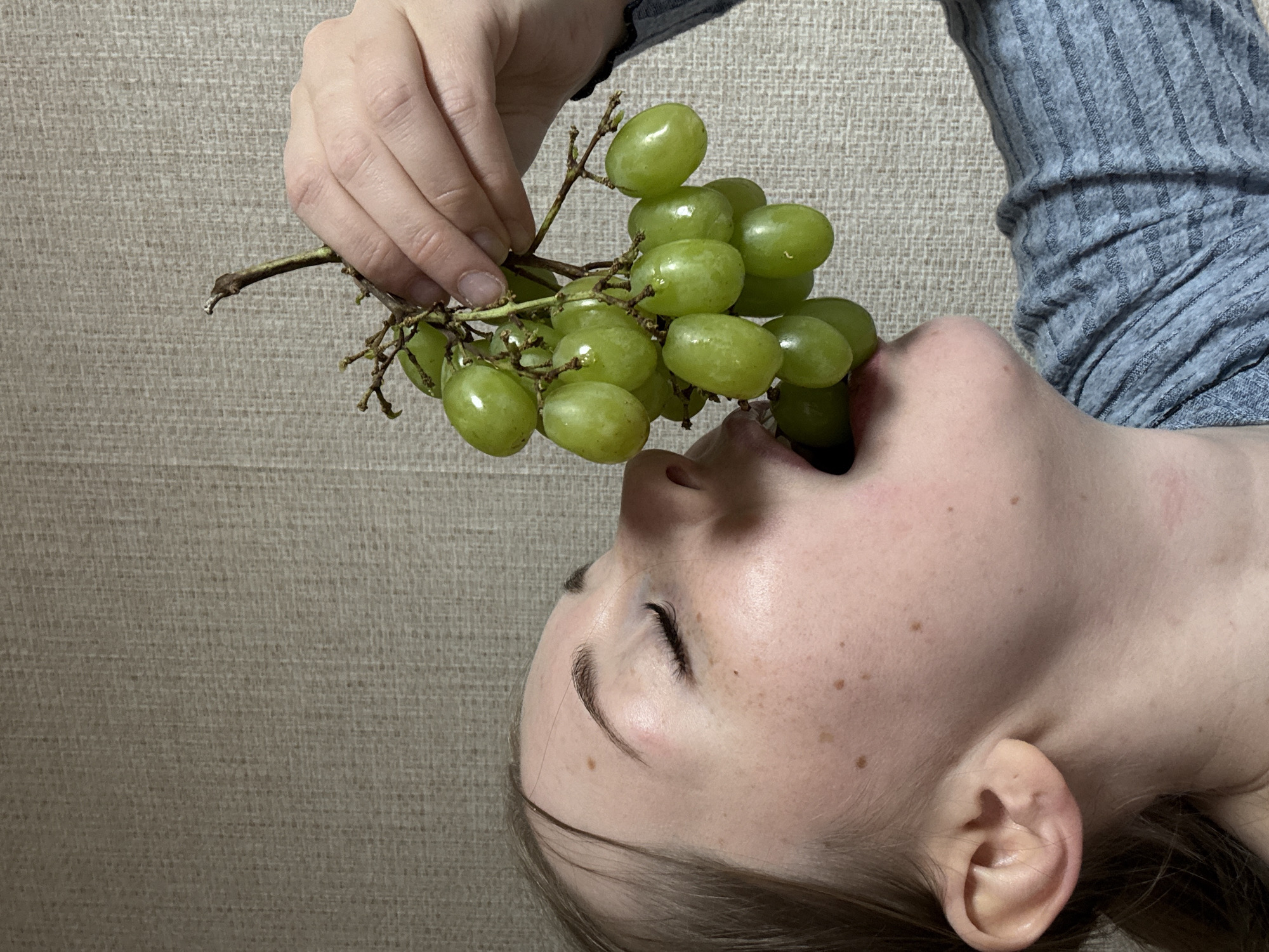 A fair-skinned person with freckles is lying on a textured beige surface. The person is wearing a grey long-sleeved shirt. The person is holding a bunch of green grapes to their mouth, with some grapes already inside.