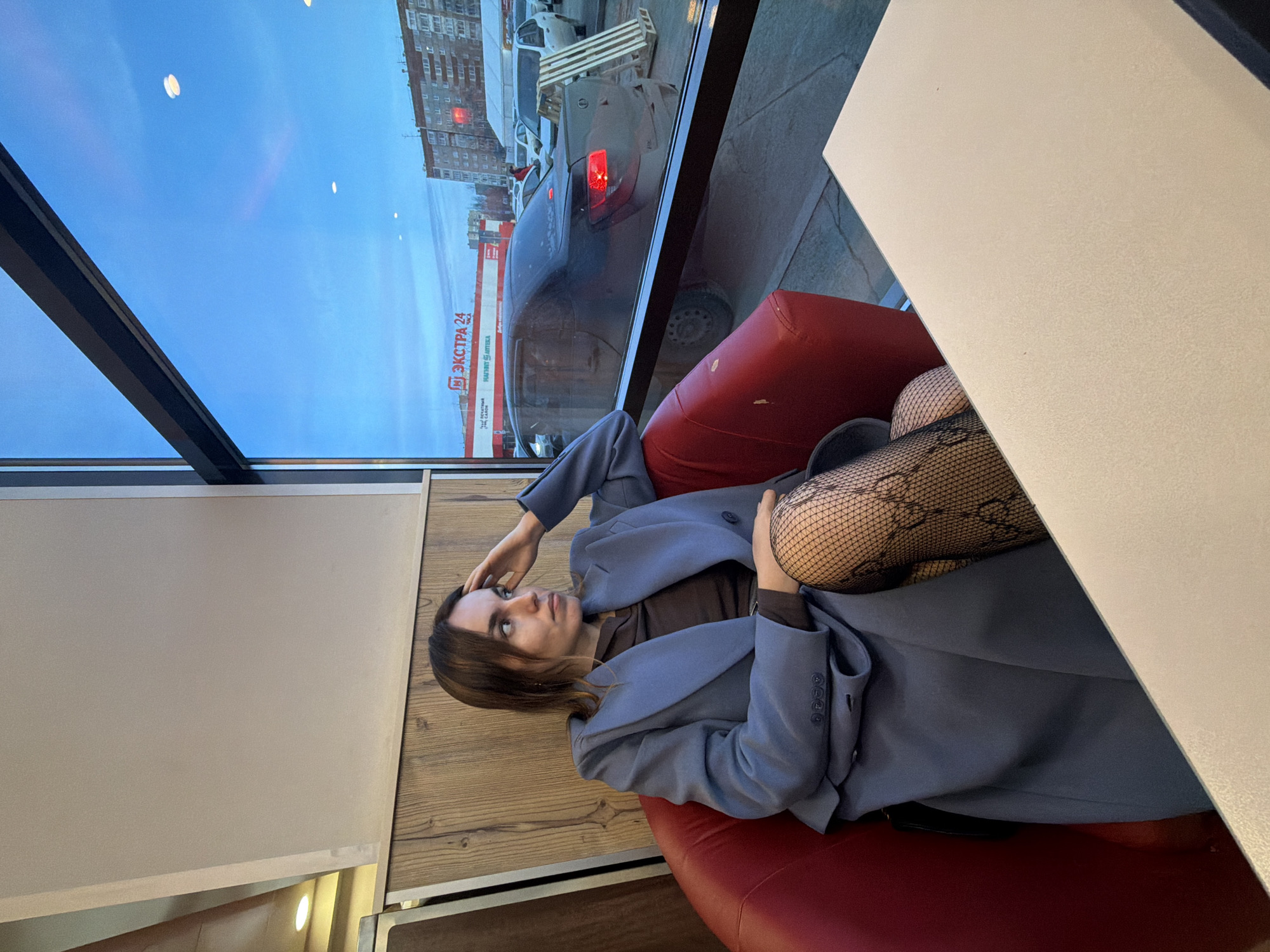 A solitary female figure is seated inside an establishment, looking out through a large window at an urban environment in Izhevsk, Russia.
The woman has medium-length brown hair and is wearing a slate-blue blazer over a dark brown long-sleeved top, paired with black patterned fishnet stockings. She is seated in a red upholstered armchair, with her left hand resting on her temple and her gaze directed towards the exterior scene visible through the window. A light-colored, rectangular table occupies the lower right foreground. The interior features vertical wooden paneling behind the woman, topped by a light beige wall section with a recessed light fixture visible to the upper left.
Outside the window, a street and parking area are visible under a pale blue sky. A dark-colored sedan, appearing dusty, is parked directly in front of the window, facing away from the viewer, with its left taillight illuminated red. Beyond it, a parking lot contains several white vehicles and a person in a red jacket. A wooden pallet leans upright near the parked cars. Across the street, a multi-story residential building is visible, and beneath it, a commercial building with a red facade displays the prominent Russian signage "ЭКСТРА 24" (EXTRA 24), with additional text "Низкие цены" (Low prices) and "НАША АПТЕКА" (OUR PHARMACY), identifying the location as Izhevsk. A white tent structure is also present in the background of the exterior scene. The overall lighting suggests dusk or an overcast day.