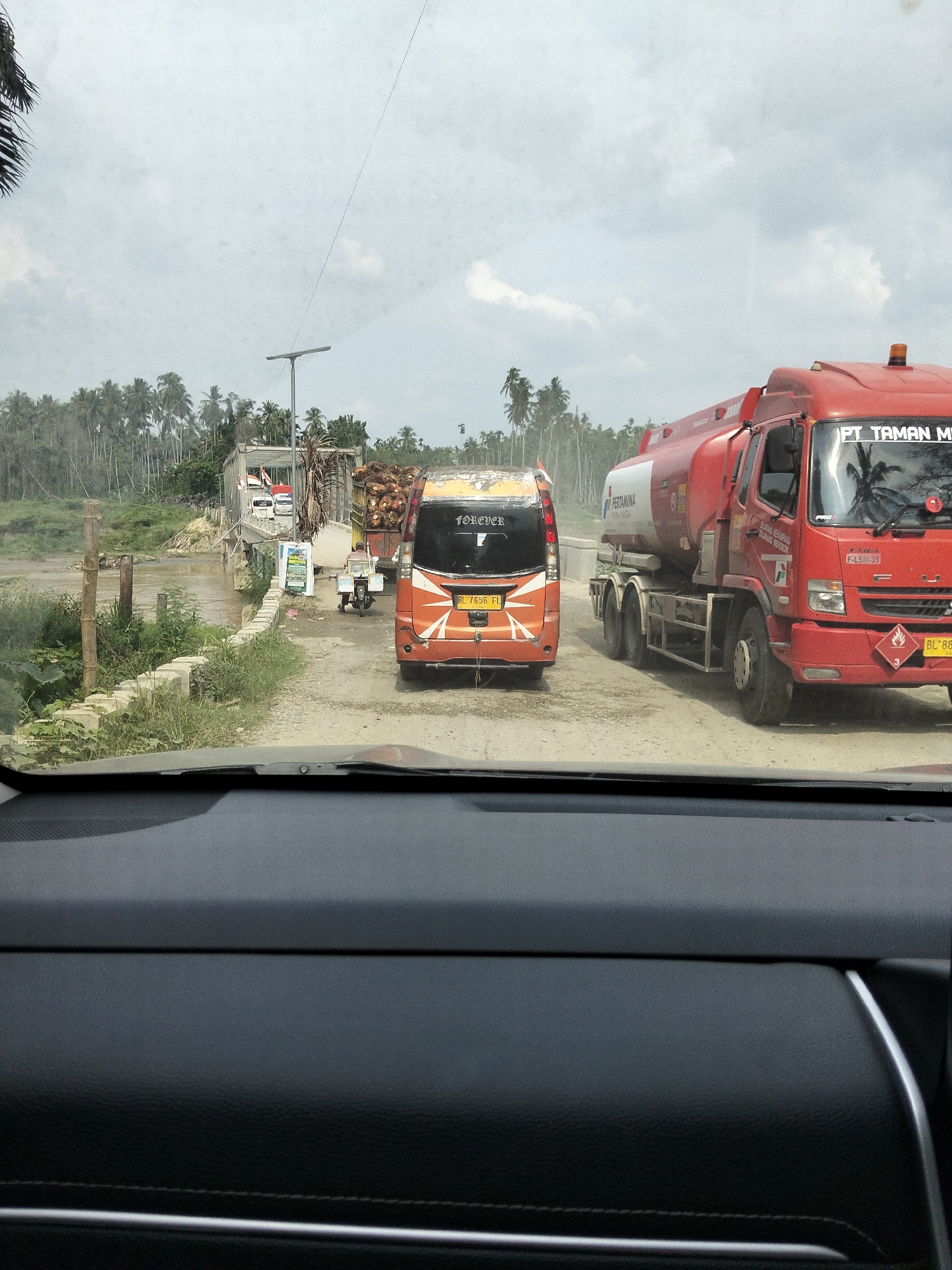 The image captures a traffic scene in Teupinreudeup, Indonesia, on a cloudy day. The foreground shows the interior of a car, suggesting the photo was taken from the driver's or passenger's seat.
The main subjects are vehicles on a dirt road. In the center, an orange minivan with the word "FOREVER" visible on its rear is stopped. To its right, a large red tanker truck with "PT TAMAN M" and "PERTAMINA" written on it is also present. The tanker truck has hazard symbols, including one with the number "3". Further back, a truck loaded with what appears to be palm tree fronds is visible near a bridge. Other vehicles and a lamppost are also present on the road.
The setting is a rural area with lush vegetation, including many palm trees, bordering the road and river. A bridge crosses the water, and the overall impression is that of a somewhat remote location. The weather appears overcast, with a dull, diffused light. The traffic suggests normal daily activity, with vehicles moving or waiting on the road. The scene is captured from a forward-facing perspective, looking out through the car's windshield. The visible text provides brand and vehicle information, such as "PERTAMINA" and license plate numbers.