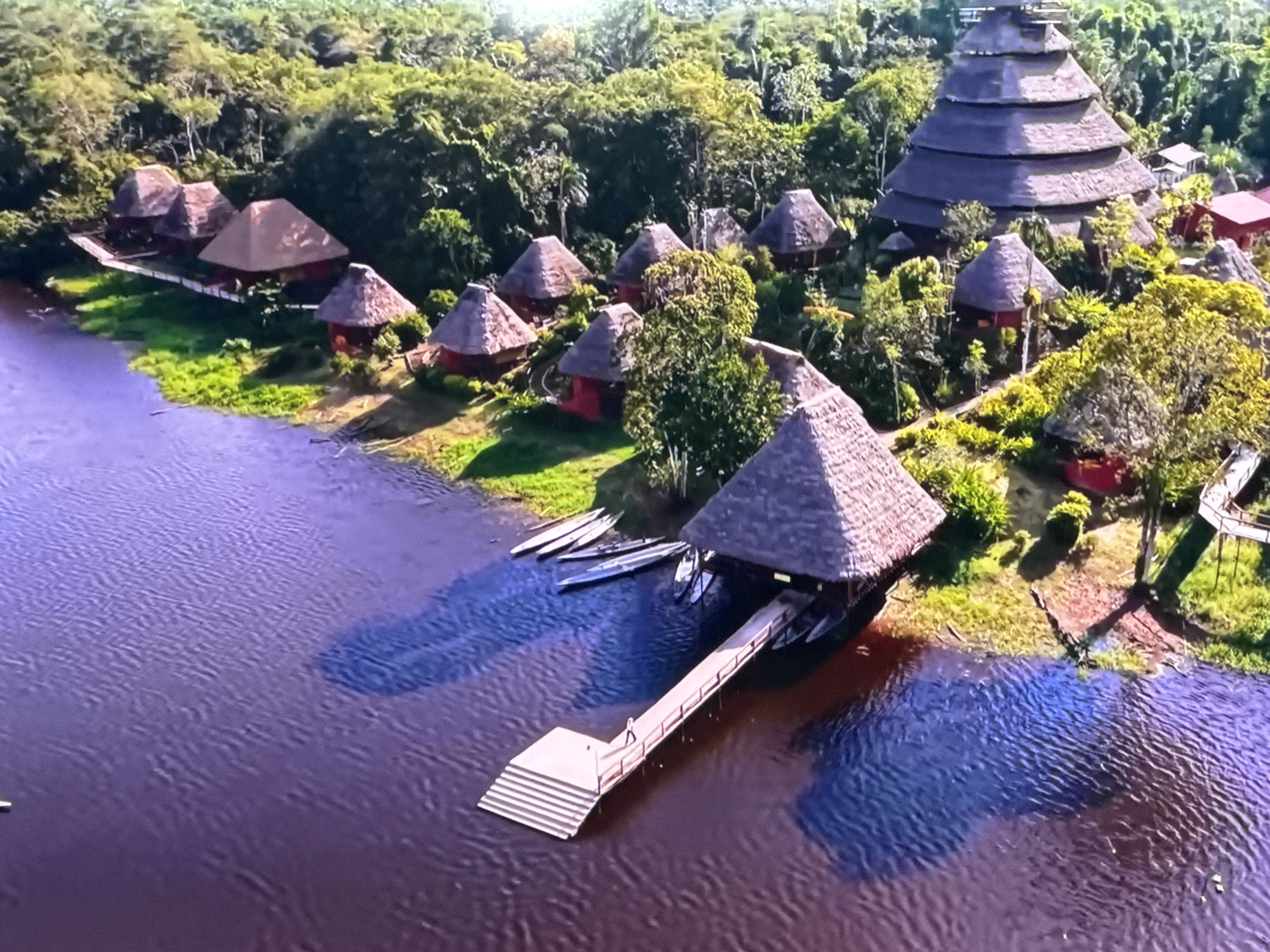This is an aerial drone shot of a jungle lodge with traditional thatched-roof huts and a larger, tiered pyramid-like structure situated along a wide, dark-water river. Several canoes are moored along the riverbank, near a wooden dock extending into the water. A single person is visible on the dock, and other structures are nestled within the dense, green foliage of the rainforest. The scene is not in Ləcət, Azerbaijan.