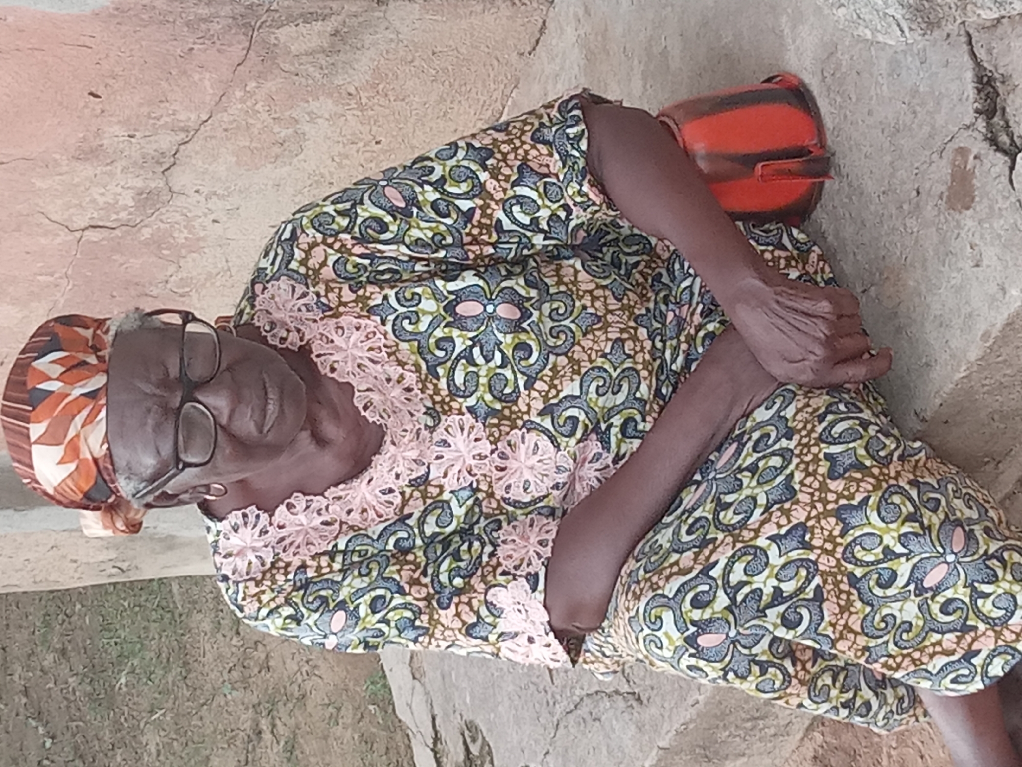 The image shows an elderly woman resting outdoors. She is wearing glasses, a headscarf, and a patterned dress with a lace collar. Her left hand rests on her lap, while her right arm is bent, with her hand near her chest. Beside her, to the upper right, is an orange and black cylindrical object, possibly a container or bag. The ground beneath her appears to be made of concrete or packed earth, with some cracks and lighter patches. The background suggests an outdoor environment with dry vegetation and possibly some rough stone structures. The lighting appears to be natural, indicating daytime, with no strong shadows suggesting midday sun. The overall scene conveys a sense of quiet repose. The location context is Sabon Tasha, Nigeria.