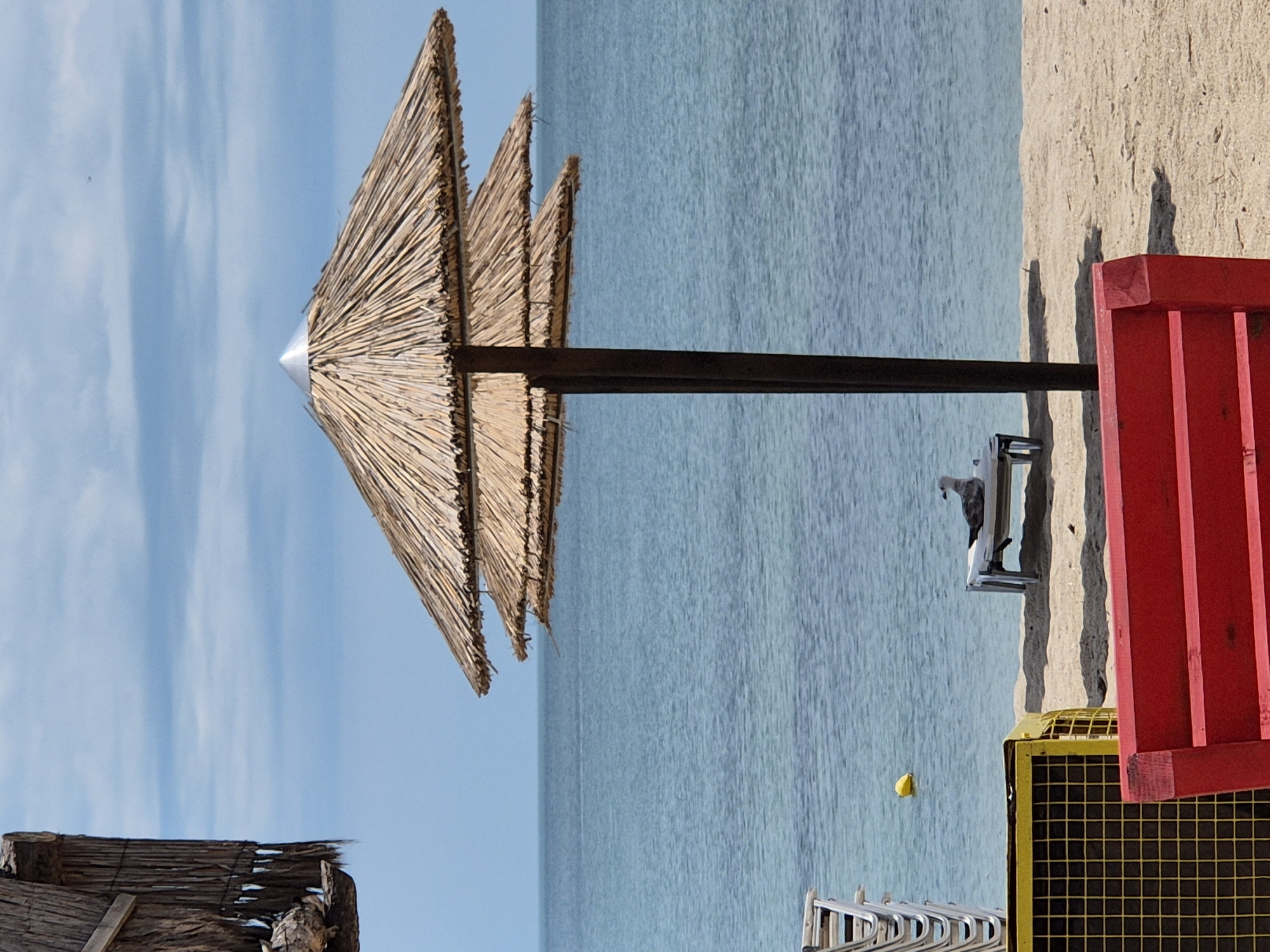 In a beach scene in an unknown city and country, a double-tiered thatched umbrella stands on a sandy beach. The umbrella has a silver cap on top. In the background, the calm, blue ocean stretches to the horizon under a partly cloudy sky. On the beach, a white lounge chair is placed on the sand, and a seagull is perched on it. To the left, a yellow metal cage and a stack of white chairs are visible. In the foreground, a portion of a red bench is shown. A yellow buoy floats in the water near the shore. The overall scene depicts a tranquil day at the beach.