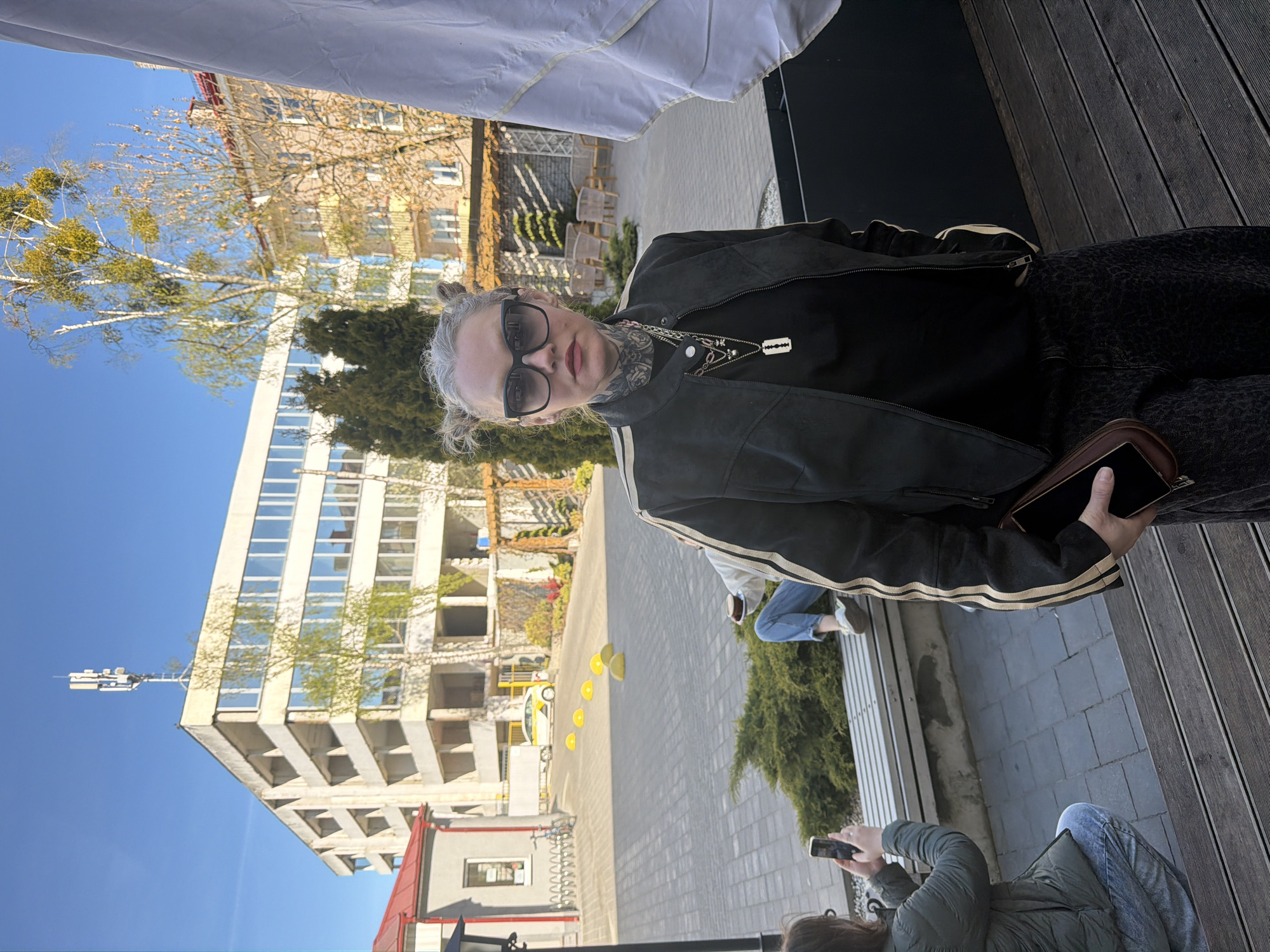 The image is taken outdoors, likely at a cafe or similar establishment, in the city of Hrodna, Belarus. A person with gray hair styled in buns and wearing sunglasses, a black jacket, and multiple necklaces, is positioned centrally in the frame. The person is holding a brown wallet and a black mobile phone. The individual is looking at the camera.
In the background, there are buildings of varying sizes and architectural styles. One appears to be an unfinished or partially demolished construction with an antenna on top. The ground is paved, and there's some greenery visible. Yellow traffic cones are lined up on the street. Other individuals are present in the image, one of whom is seated and using a mobile phone.