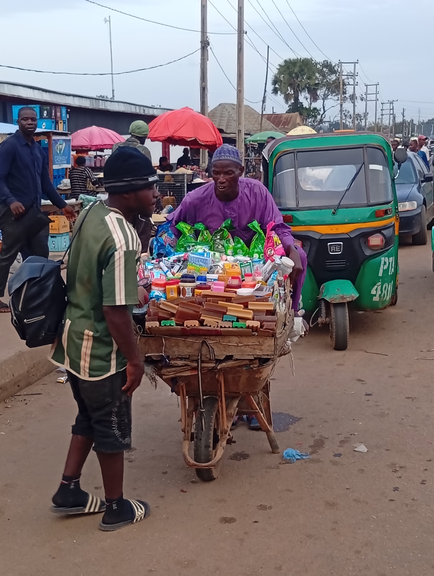 In Bukuru, Nigeria, a street scene unfolds with a man in a purple robe standing behind a cart laden with packaged goods, possibly soaps, candies, and toiletries, next to a green and yellow motorized rickshaw. Another man, wearing a green and white striped shirt and shorts, stands to the left, a black backpack slung over his shoulder. They appear to be interacting or engaged in a transaction. The environment suggests a busy market or roadside area, with other people, stalls, and vehicles, including a dark-colored car, visible in the background. The sky is overcast.