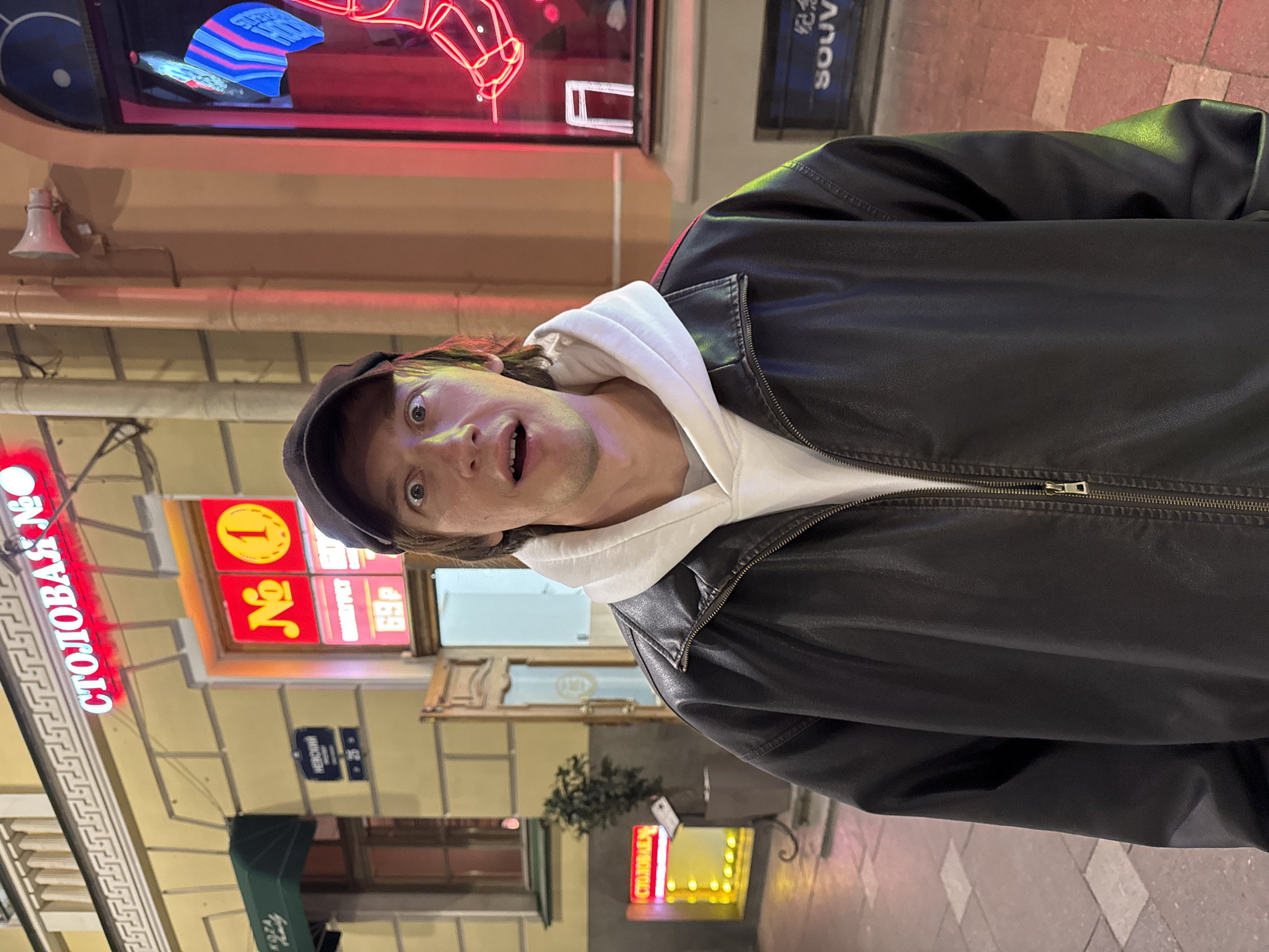 A man is standing on Nevsky Prospekt in Saint Petersburg, Russia. He is wearing a black leather jacket over a white hoodie and a black baseball cap. He is looking at the camera with a surprised expression. In the background is Столовая №1, and the address Nevsky 26. The buildings are brightly lit at night.