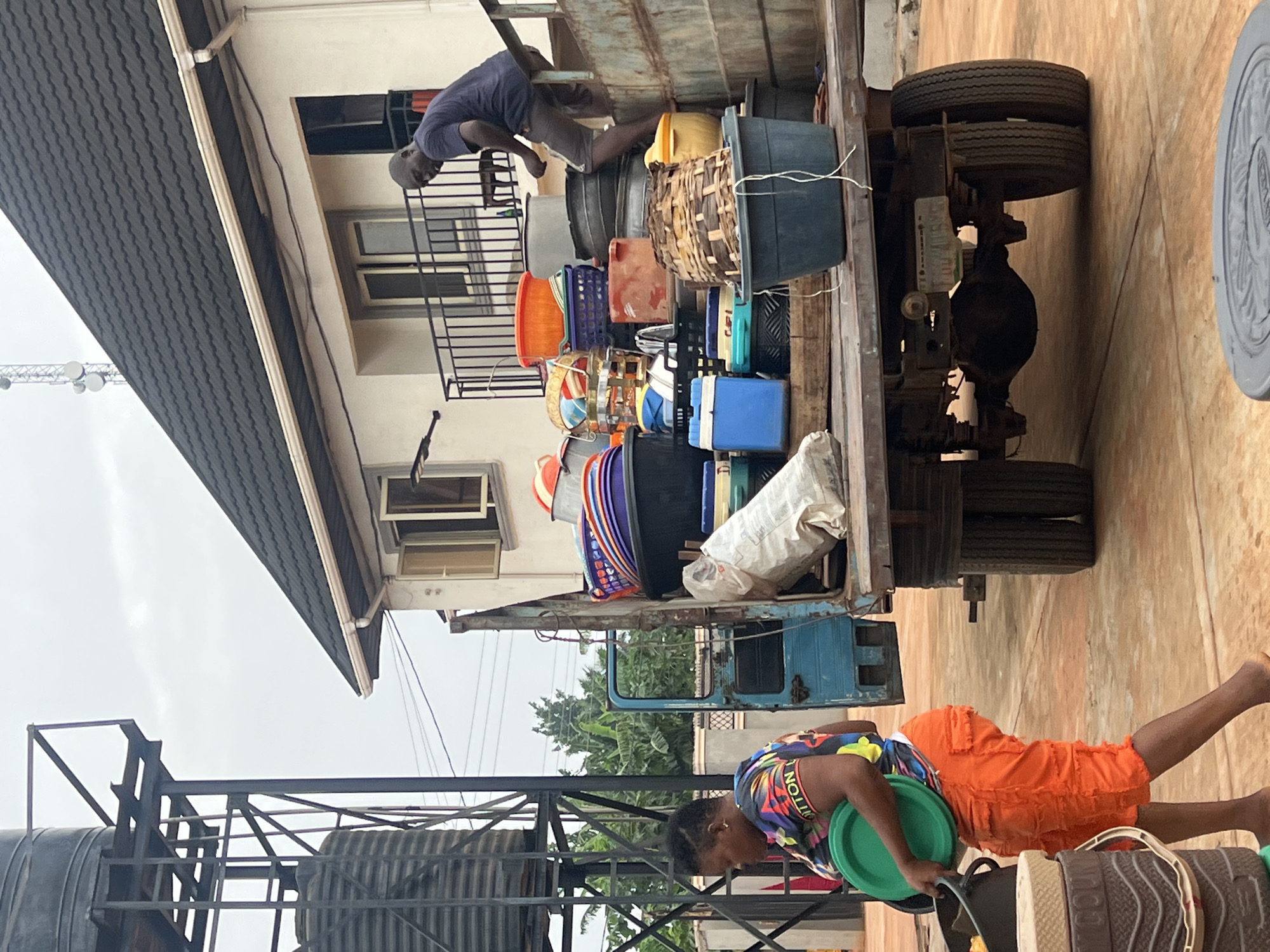 The image captures an outdoor scene in Onitsha, Nigeria, during an overcast day. A light blue, flatbed truck is heavily loaded with various goods and positioned next to a two-story building.
On the truck bed, a man in a dark blue t-shirt and shorts is actively arranging or loading numerous items. These include a variety of colorful plastic containers, such as large black tubs, yellow basins, orange and purple baskets, several woven brown baskets, and white sacks. The rear wheels and axle of the truck are clearly visible.
In the lower foreground, another individual stands, wearing a brightly patterned, multi-colored shirt that reads "VUITTON" and vibrant orange cargo shorts. This person is holding a green plastic basin over a dark container, engaged in handling goods. Nearby on the ground are several patterned mats or baskets, one of which shows partially visible text "INCO".
The background includes a cream-colored building with dark-framed windows and a balcony railing, topped by a dark, ridged roof with a white gutter. To the left, a tall metallic structure, possibly a water tower or communication mast, stands against the cloudy sky. Another large, corrugated water tank structure is visible in the lower left. The ground is a light, paved surface. The scene depicts a busy commercial or logistical activity involving the transport and handling of goods.