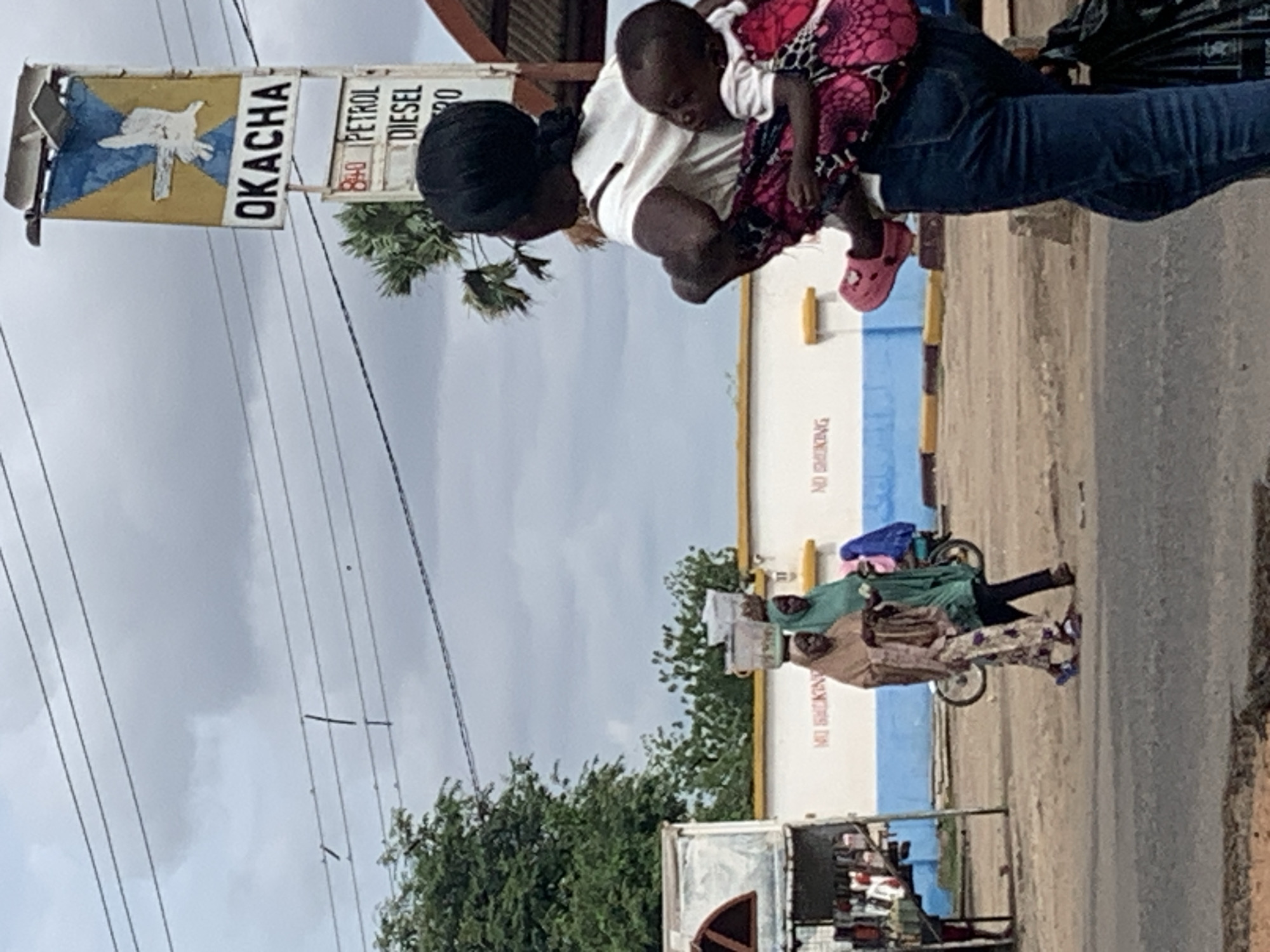 This image captures a street scene in Shendam, Nigeria, under an overcast sky. In the foreground, positioned on the right, a woman with dark hair, wearing a white shirt and blue jeans, walks away from the viewer, looking slightly towards the right. A baby, wearing a white shirt and pink Crocs, is secured to her back with a pink and black patterned fabric carrier, its head resting on the woman's right shoulder. The woman carries a black bag in her left hand.
To the left of the foreground woman, in the midground, two women are walking from left to right on an unpaved surface adjacent to a paved road. The lead woman wears a light brown head covering and a patterned dress, balancing a translucent container on her head and holding an item in her hands. The woman behind her wears a green head covering and a green top, also holding an object. Behind them, a light-colored wall with a blue stripe along its base displays repeating red text that appears to be "NO SMOKING." A bicycle is partially visible behind the second woman.
Further into the background, on the right, a tall roadside sign for "OKACHA" is prominent. The sign features a yellow background with a blue 'X' motif and a white bird or dove-like figure in the center. Below "OKACHA," a smaller sign indicates "PETROL" and "DIESEL," with "840" visible above "PETROL," likely denoting fuel prices. Electrical power lines stretch across the cloudy sky above the street. Green foliage from trees is visible on both sides of the OKACHA sign and in the far-left background. On the far left, a makeshift stall or small shop, appearing to sell various goods, stands by the roadside, with a person's leg partially visible near it. The ground consists of a dark paved road and lighter unpaved sections.