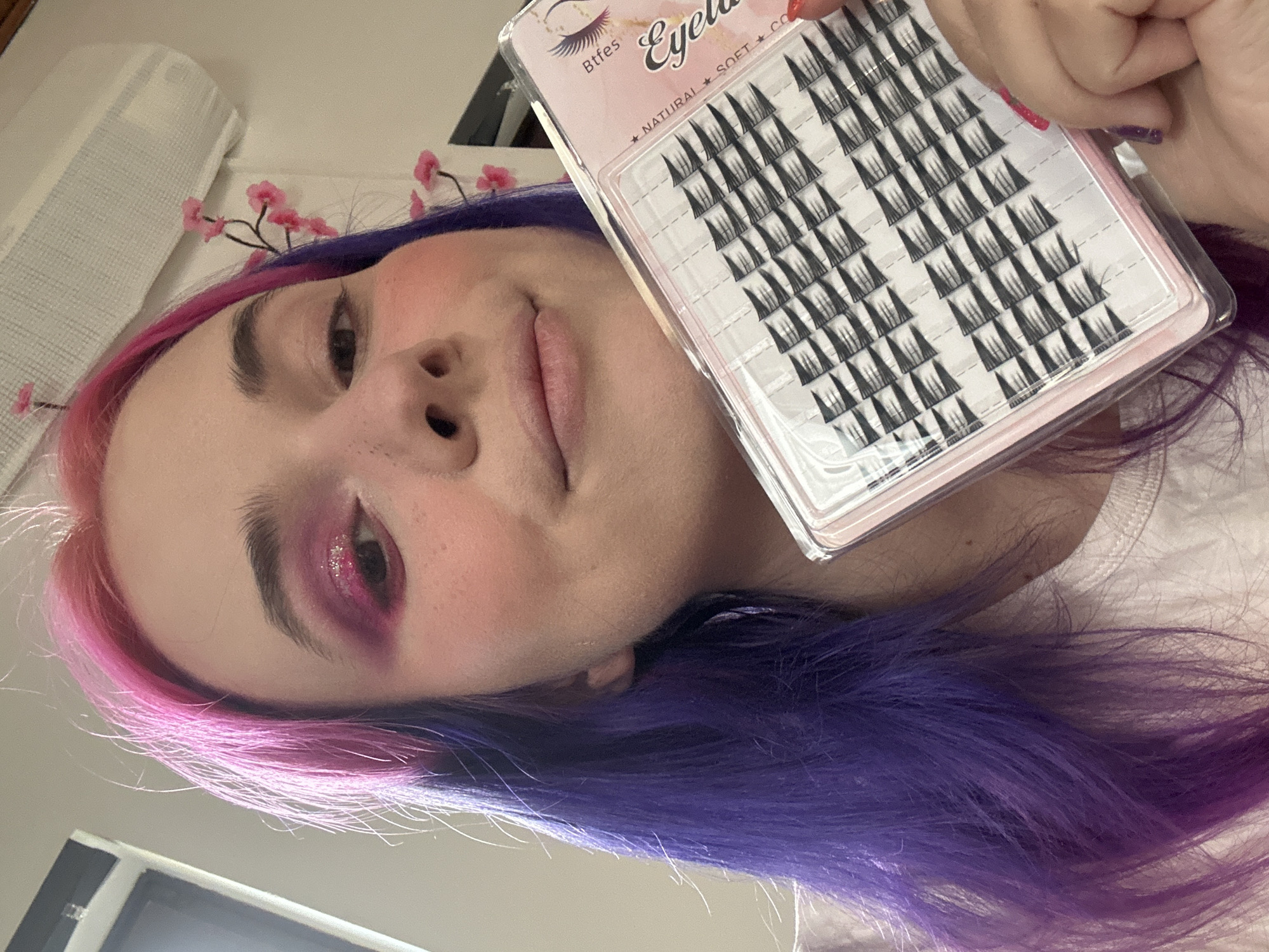 A woman with pink and purple dyed hair is holding a package of false eyelashes in her right hand. She is looking at the camera with a slight smile. Her makeup includes pink eyeshadow and blush. She has a small decorative sprig of pink flowers attached to her hair. She is wearing a white shirt. The background is a white wall. There is no evidence that this image was taken in the city of Zagreb, Croatia.