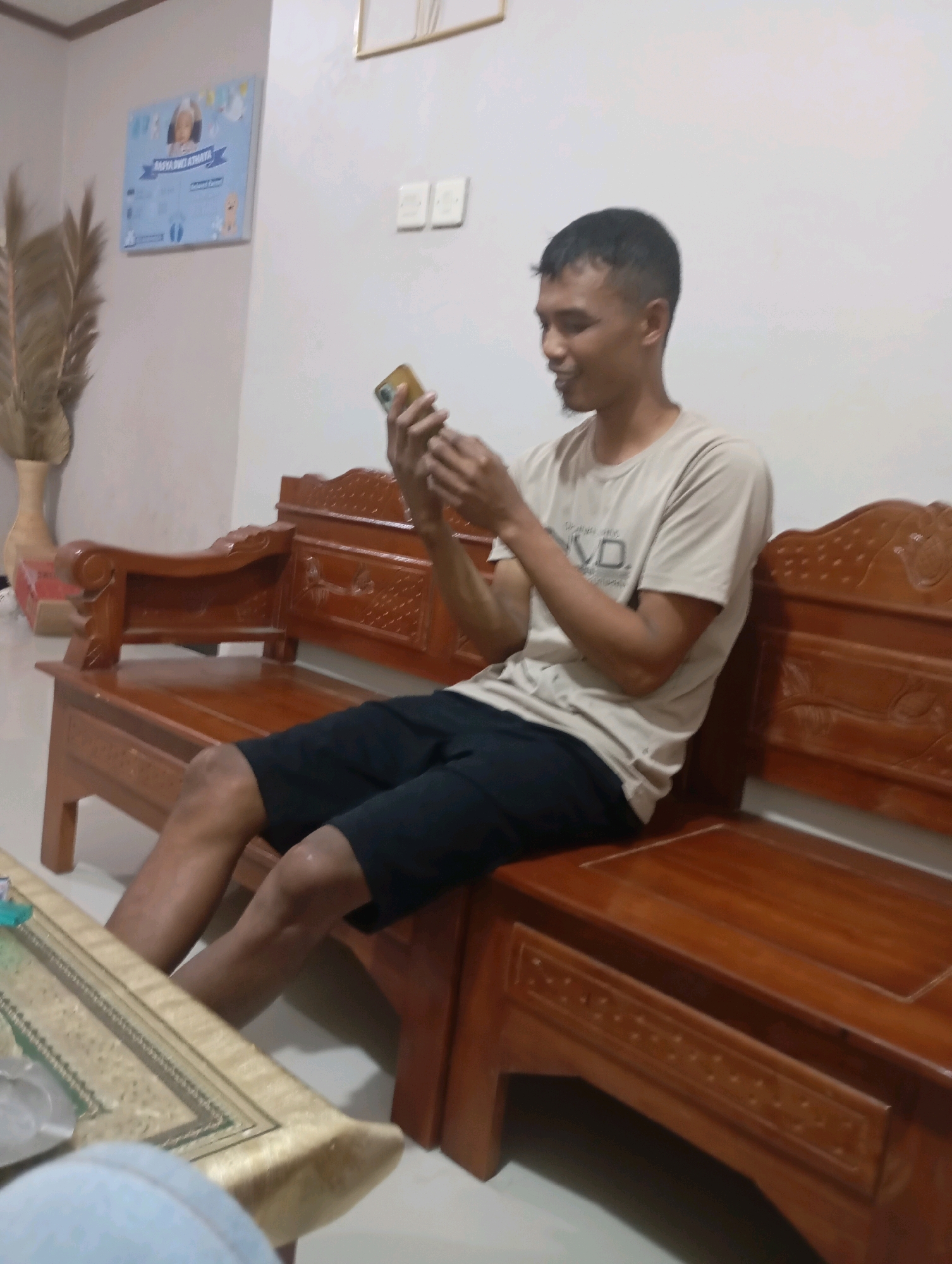 A man is sitting on a wooden bench in a room, holding and looking at a mobile phone. He is wearing a light-colored t-shirt and black shorts. The bench is intricately carved and appears to be made of dark wood. In the background, there is a light-colored wall with two light switches and a framed picture of a baby. To the left of the picture, a dried palm frond is displayed in a vase. A table with a patterned cloth is in the foreground. The scene is indoors, likely in a residential setting in Ketepung, Indonesia.