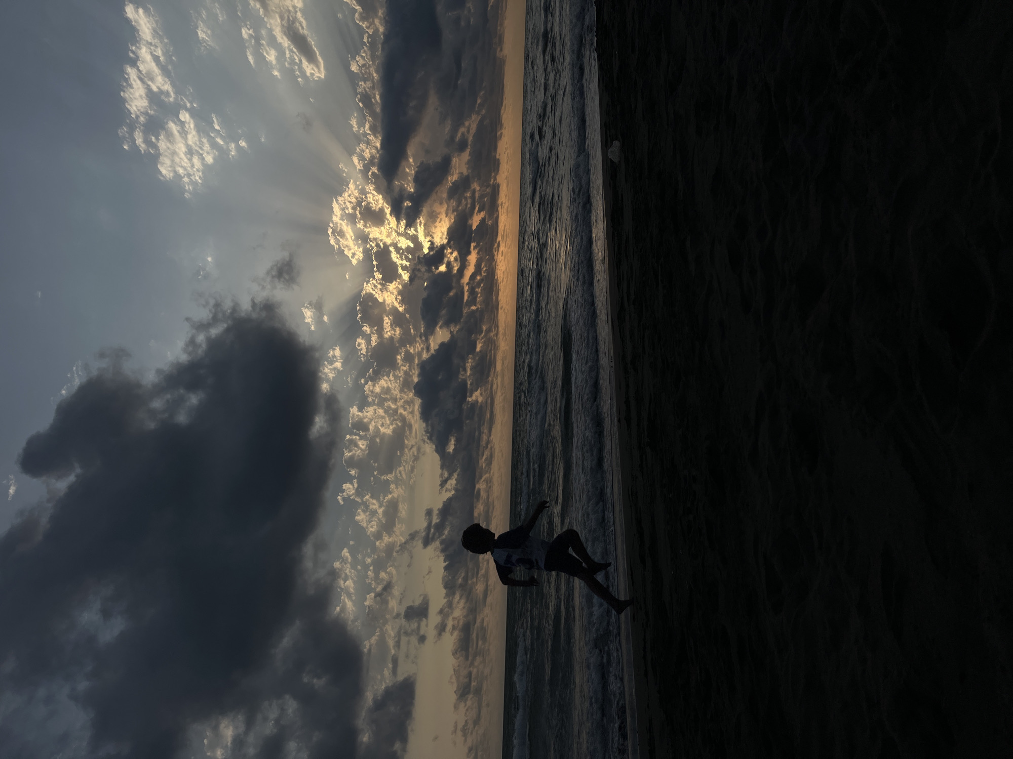 A silhouetted child runs along a dark sand beach towards the ocean at sunset. The sky is dramatic, with dark, heavy clouds on the left and sun rays bursting through lighter clouds on the right. The ocean is choppy with visible waves. The sand in the foreground is dark and appears textured. The location is likely the coast of the Black Sea near the village of Supsa in Georgia.