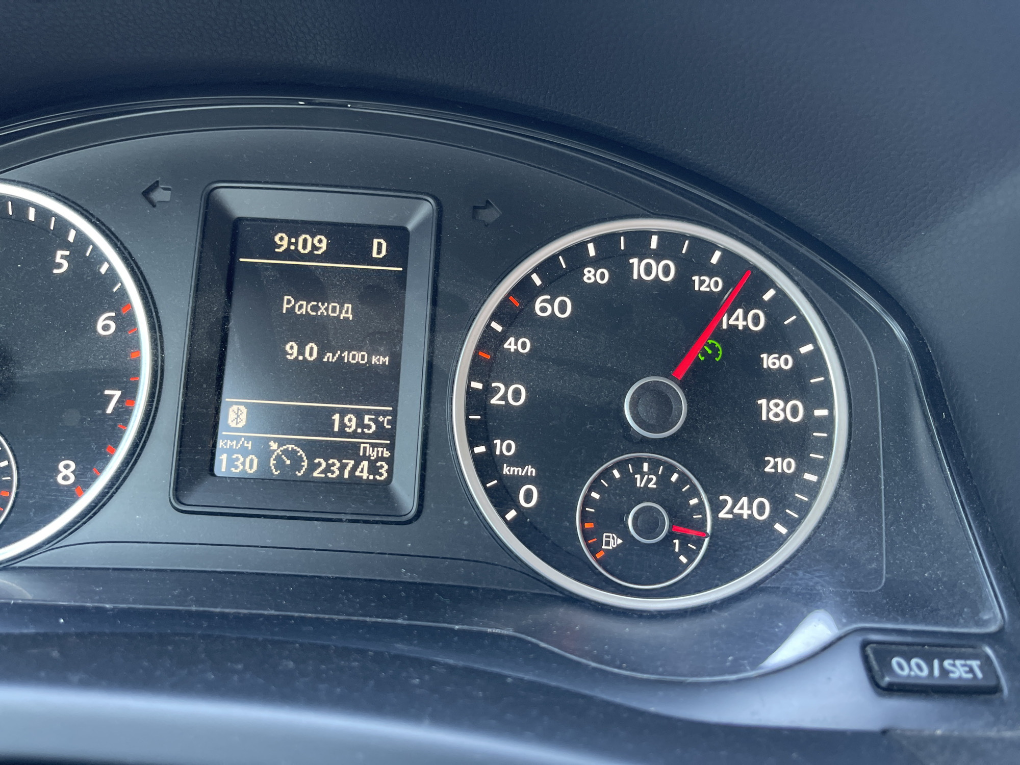 The image shows a close-up of a car's dashboard. The dashboard displays a digital clock showing 9:09, the gear selector is in "D" (Drive), and the fuel consumption is 9.0 liters per 100 kilometers. The outside temperature is 19.5 degrees Celsius. The odometer shows 2374.3 kilometers traveled. The speedometer indicates the car is traveling at 130 km/h. A fuel gauge shows the fuel level is slightly above the "1" mark. The image is taken from inside the car, looking at the instruments. The environment appears to be daytime, with natural light reflecting off the dashboard. There is no visible indication of the location being Hrozyne, Ukraine.