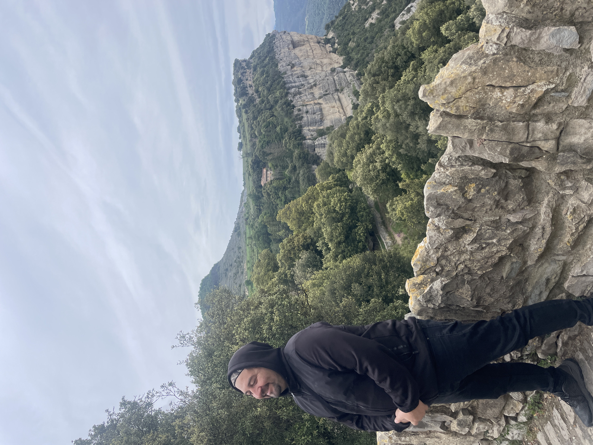 A man stands on a stone ledge in Tavertet, Spain. He is wearing a black hooded jacket, black pants, and black shoes. He is looking down and smiling. The background features a mountainous landscape with trees and a cloudy sky. A prominent rock formation is visible in the distance. The foreground consists of the stone ledge and the surrounding vegetation. The photograph appears to have been taken from a slightly elevated position.
