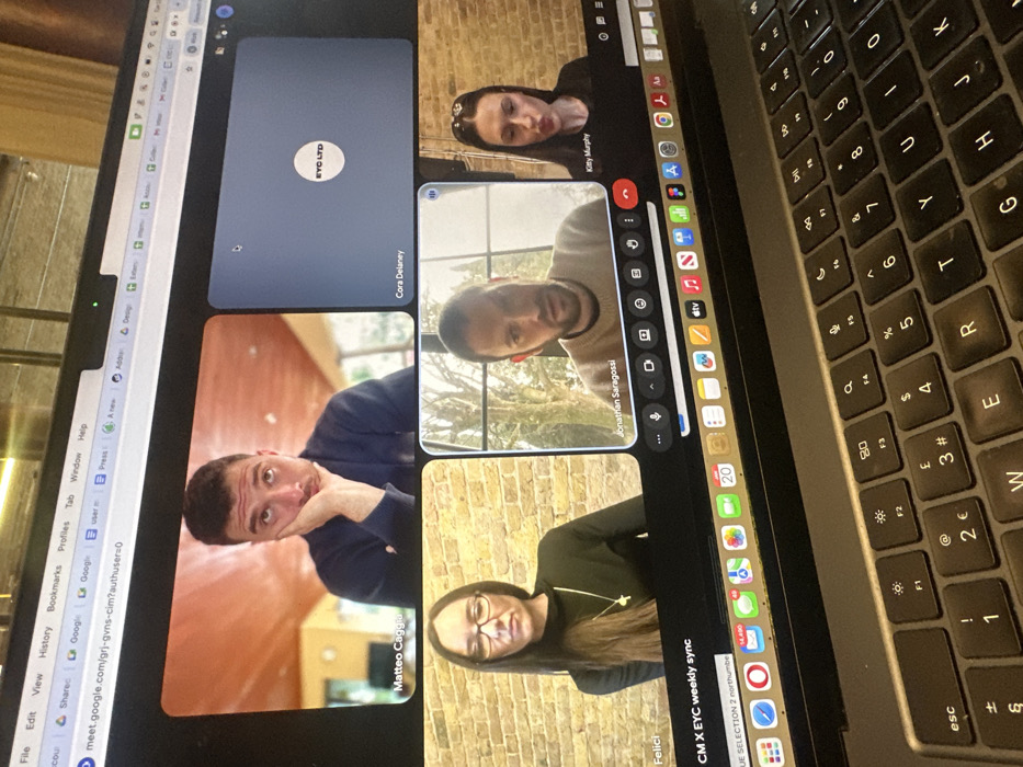 A close-up, elevated view captures a silver laptop screen displaying a Google Meet video conference, angled towards the viewer. The screen shows five participants in a grid layout. In the top-left quadrant, Matteo Caggia, a man with short dark hair, rests his chin on his left hand, looking upwards with a seemingly bored or tired expression against a background featuring a wooden ceiling and light-colored wall. The top-right quadrant shows a dark blue panel with a white "EYC LTD" logo and the name "Cora Delaney," indicating no video feed. The bottom-left quadrant displays Felici, a woman with long dark hair and glasses, wearing a black top with a cross pendant, seated in front of an exposed brick wall. In the bottom-middle quadrant, Jonathan Saragossi, a man with a beard, is visible against a bright background with trees outside a window, appearing to speak. The bottom-right quadrant features Kitty Murphy, a woman with dark hair and hair clips, with pursed lips, also against an exposed brick wall. The meeting title "CM X EYC weekly sync" is visible at the bottom of the video conference interface. Below the screen, the laptop's QWERTY keyboard with a "£" currency symbol is partially visible, indicating a UK layout. The macOS dock is visible at the bottom of the screen, displaying numerous application icons and a calendar icon showing "20." The browser's address bar displays "meet.google.com" with multiple open tabs. The scene depicts a virtual business meeting taking place on a laptop, in London, United Kingdom.