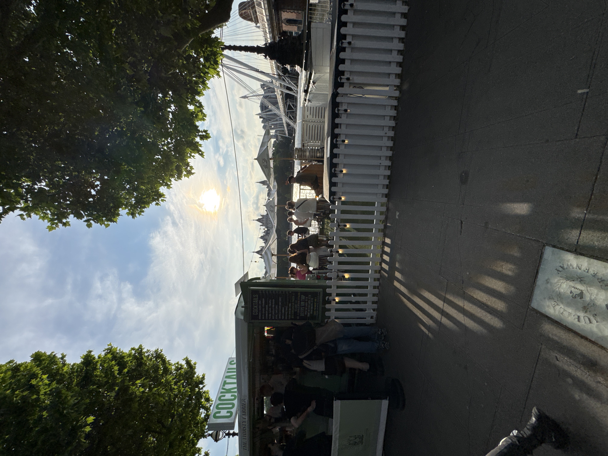 The image depicts a scene along the South Bank of the River Thames in London, United Kingdom, with the Golden Jubilee Bridges in the background. A temporary outdoor bar, "The Thirsty Ferrier," is set up, featuring a prominent "COCKTAILS" sign. People are gathered at the bar and seated at tables, some under umbrellas, enjoying drinks. The sun is setting, casting long shadows across the paved walkway and creating a warm glow in the sky. The overall atmosphere suggests a leisurely evening outdoors.