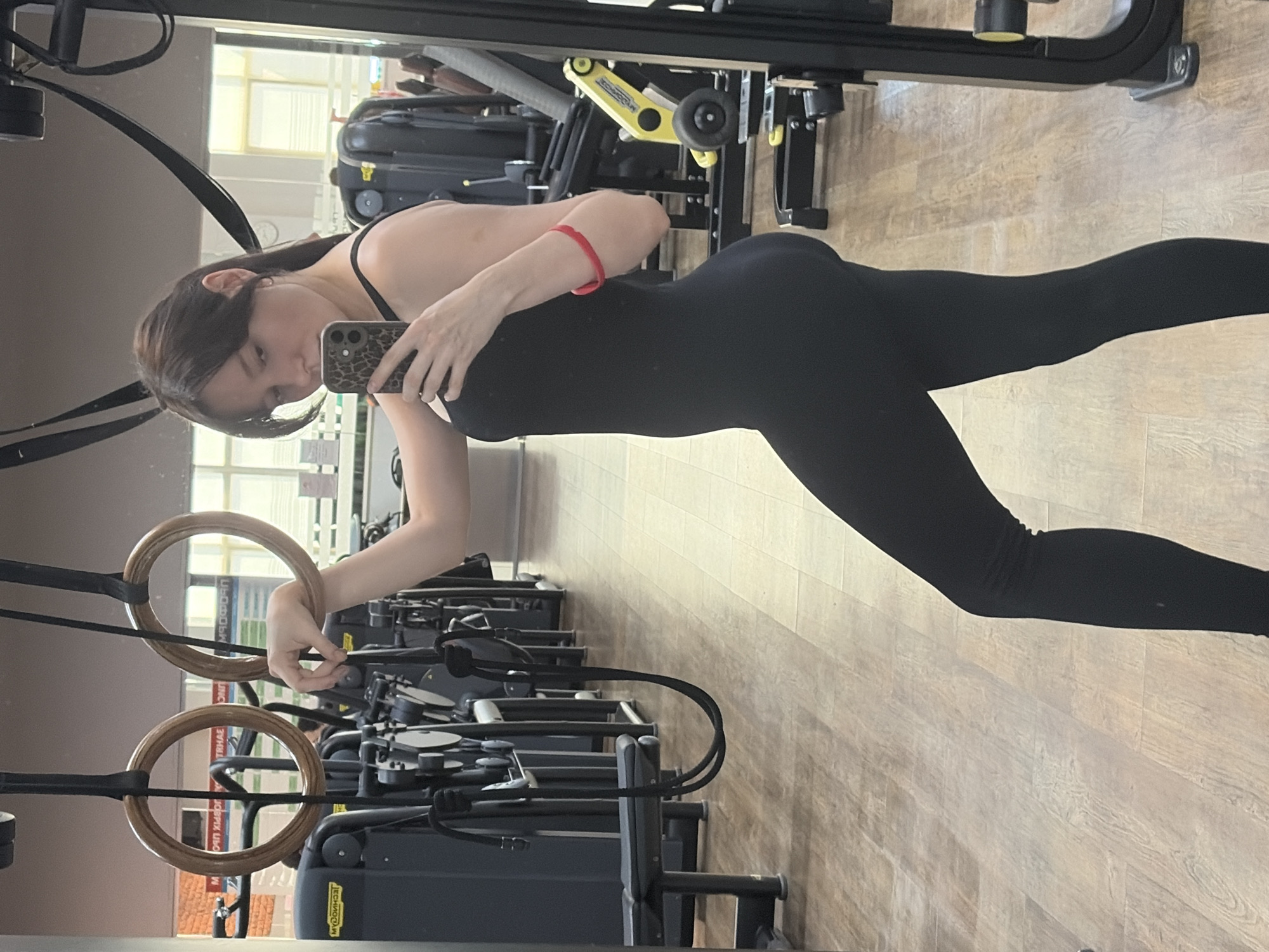 A young woman with dark hair and bangs is taking a mirror selfie in a gym. She is wearing a black leotard and black leggings. She has a red bracelet on her right wrist. In the background, there are gym machines and wooden flooring. The location is the "Dynamo" sports complex in Chaikovsky, Russia, identified by visible Russian text on gym equipment in the background.