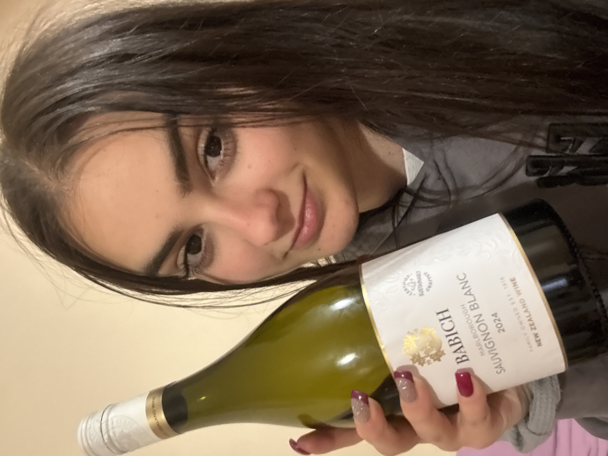 A young woman with dark hair is holding a bottle of Babich Marlborough Sauvignon Blanc 2024 New Zealand Wine. The bottle is green with a white label featuring gold accents. The woman is smiling slightly and her nails are painted with a two-toned design of burgundy and silver glitter. The image appears to be taken indoors, and there is a glimpse of a gray hoodie on the woman's shoulder. There is no indication of the location in Blagoevgrad, Bulgaria or other details.