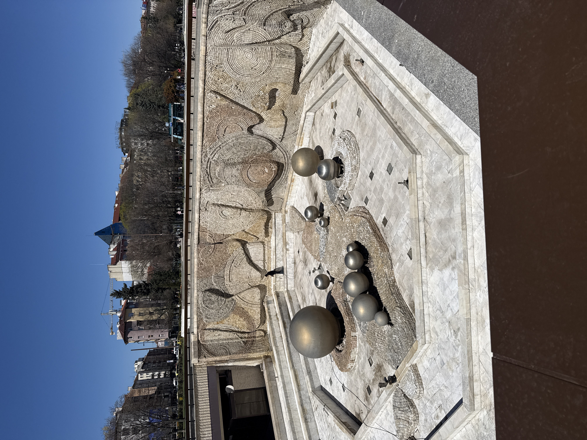 This is a high-angle, eye-level shot of the monument in Sofia, Bulgaria. The camera is oriented perpendicular to the ground, looking down and slightly to the side. The fountain, with its mosaic wall and metallic spheres of various sizes, dominates the foreground. The wall is patterned with concentric circles. A nearby building can be seen through an opening in the fountain structure. Beyond the monument, a city skyline is visible under a clear blue sky. Buildings of varying heights and architectural styles, interspersed with trees, line the horizon. A crane is visible in the distance, indicating ongoing construction. The overall impression is one of urban landscape combined with artistic monumentality.
