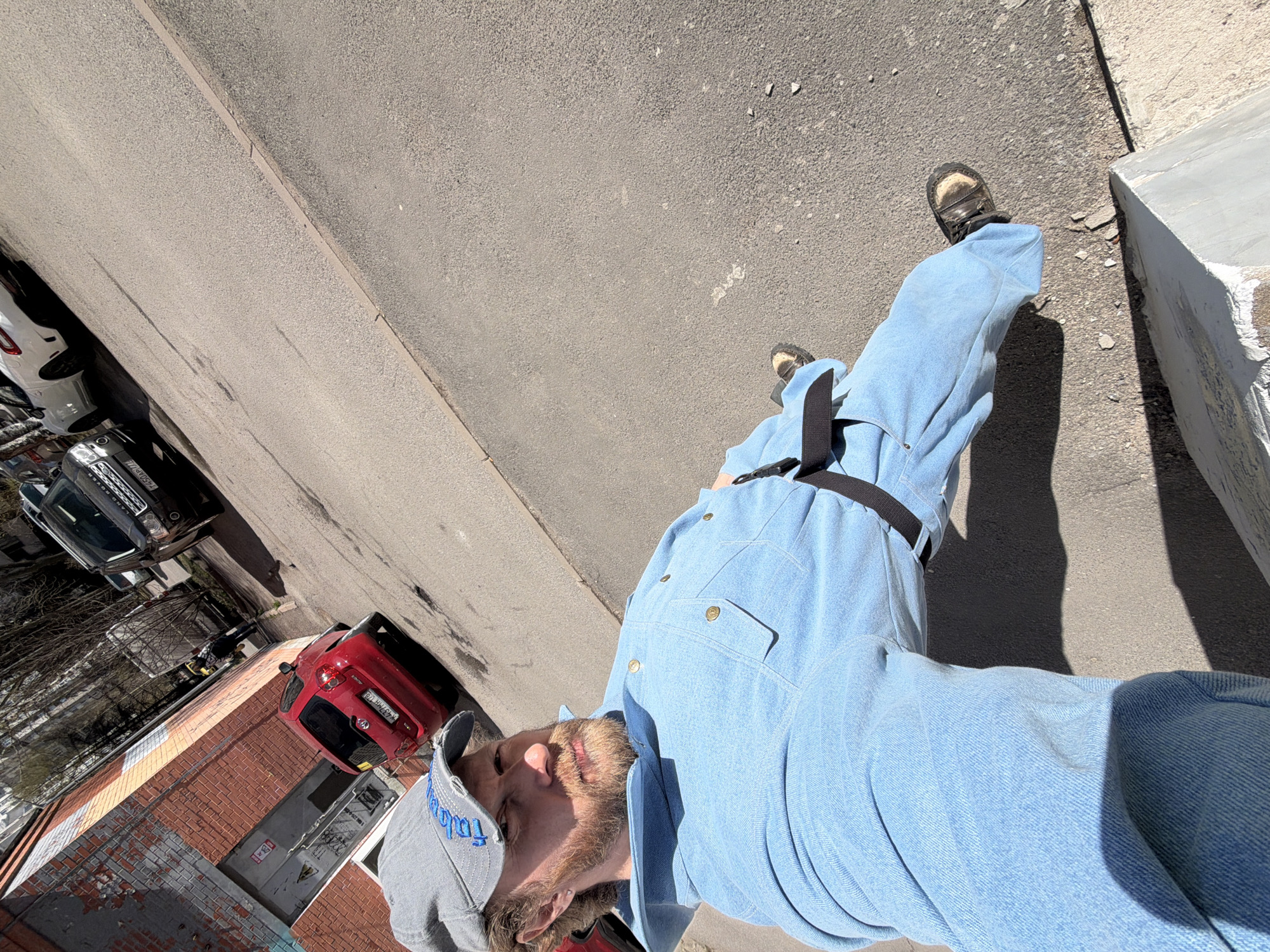 An outdoor, vertically rotated photograph captures a man and an urban street scene in Saint Petersburg, Russia. The man, positioned in the foreground and appearing to lie horizontally due to the camera's tilt, has a beard and is wearing a light blue denim-like jumpsuit or overalls with a black belt, dark sandals, and a grey cap embroidered with blue "Toblerone" text.
The background shows an asphalt road extending diagonally, with several parked cars. To the left, a black Land Rover SUV is visible with a partial license plate "H543 HM 178". Further down the street, a white car is partially seen. To the right, a red hatchback, possibly a Toyota Yaris, is parked by a brick building. Its license plate, "A311 HM 178", further indicates the Saint Petersburg region (code 178).
The scene is brightly lit by the sun, casting distinct shadows, suggesting mid-day to afternoon. The brick building appears weathered, with signs of grime or damage on its lower sections, and a dark doorway is visible. A yellow sign with an illegible symbol (possibly "STOP") is affixed to the door. A large, rough concrete block stands to the right of the man's feet. The overall setting is a typical city street or alleyway.