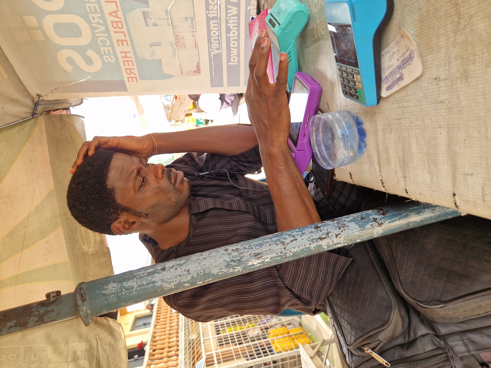 A Black man, appearing to be in his late 30s to early 40s, is seated at a table under a striped awning. He is wearing a dark brown, pinstriped tunic. His right hand is on his forehead, and his gaze is directed downwards, suggesting contemplation or fatigue. The table surface is covered with a rough, beige cloth and displays several items: a purple point-of-sale (POS) terminal, a teal POS terminal, a clear plastic cup filled with water, and a small handwritten note. In the background, a metal pole supporting the awning is visible, as are other structures and people, suggesting an outdoor market or street vendor setting. The overall scene implies a business transaction or operational context, possibly in a commercial area of Kabong, Nigeria.