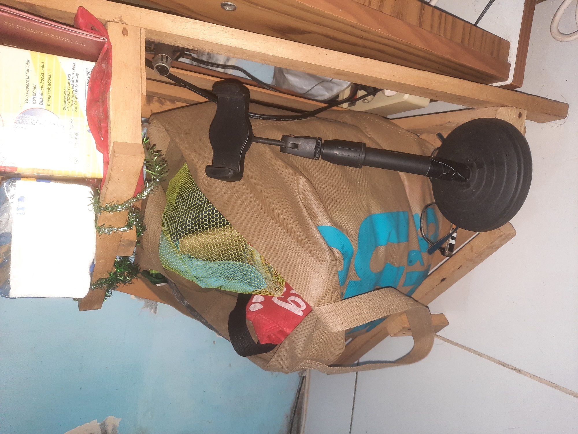 The image depicts a close-up, angled view of a wooden shelf unit supporting various items. On the top shelf, a white object, possibly a tissue box or a food item packaging, is visible. Green tinsel is draped over the front of the shelf. Below this, a large, brown canvas bag with blue lettering is the central focus. Inside the bag, a bright red object with white lettering, resembling a child's toy or game piece, is partially visible, along with a yellow mesh bag. To the right of the canvas bag, a black microphone stand with a phone holder attachment is positioned. The background shows a blue wall and parts of the wooden shelf structure, including visible screws and wiring. The scene appears to be indoors, possibly in a home or storage area, with the floor in the foreground being a light-colored tiled surface with a visible grout line. The overall impression is one of clutter and everyday items. The image does not contain any information that would allow for the determination of its location in West Jakarta, Indonesia.