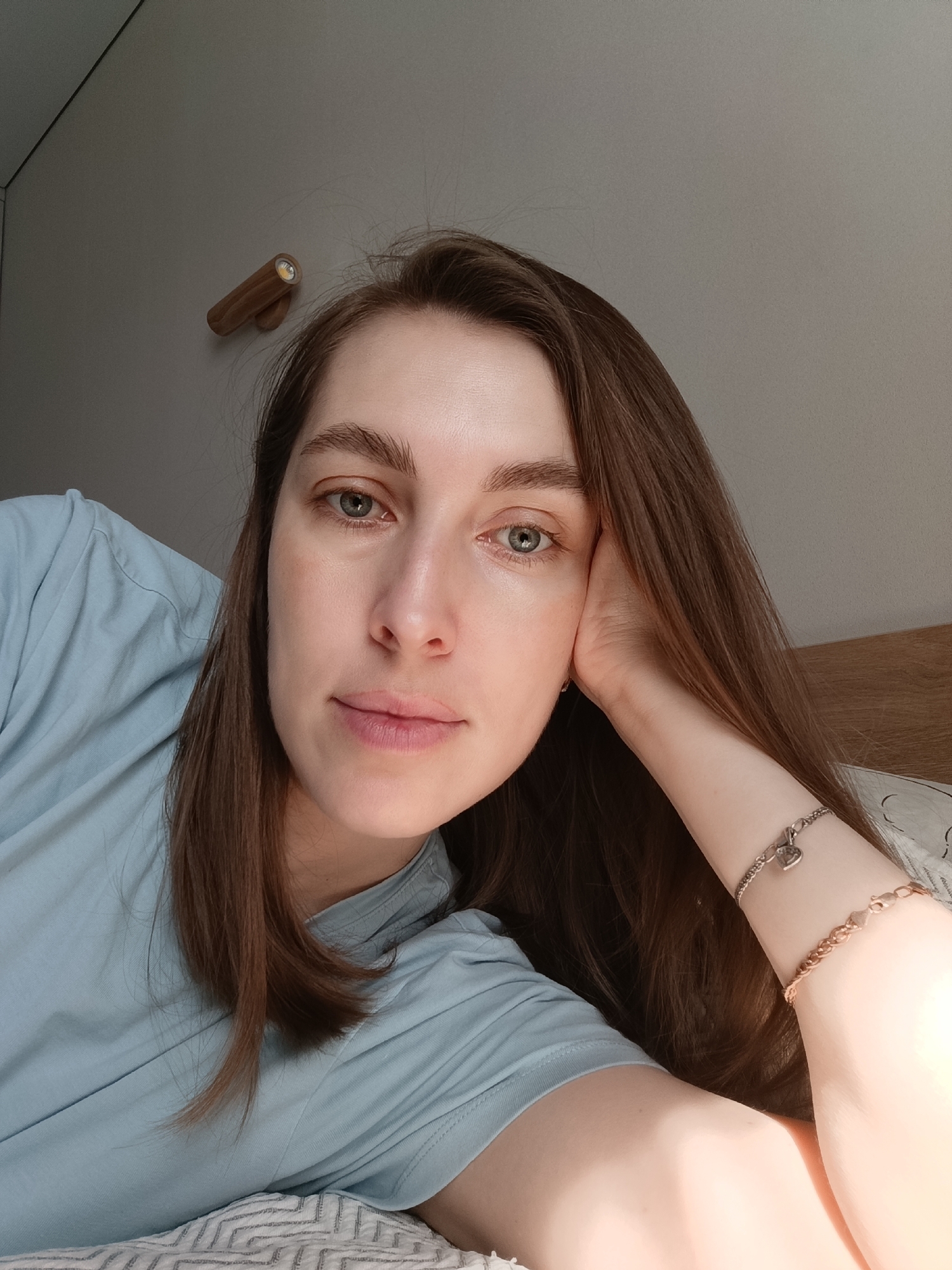 A fair-skinned woman with long, straight, brown hair and light blue eyes is lying down. She is wearing a light blue t-shirt. Her right hand is supporting her head. Two bracelets adorn her right wrist. The background shows a neutral-colored wall with a wooden wall light. A portion of the headboard is visible behind her head. The bedding has a patterned, textured surface. No evidence supports a location in Chaikovsky, Russia.