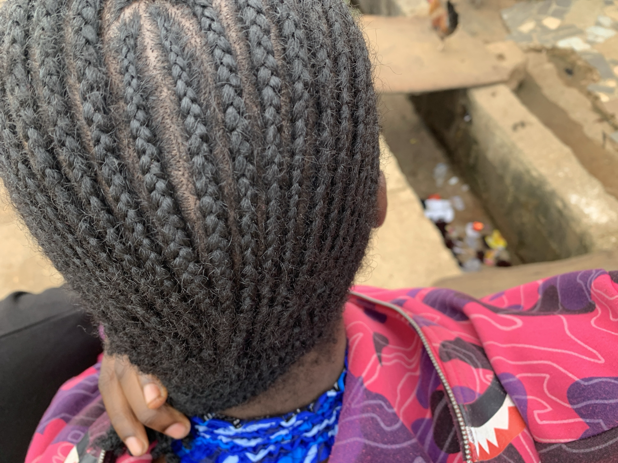 An overhead, close-up photograph depicts the upper torso and head of an individual with dark skin, positioned outdoors in Rafin Par, Nigeria. The subject's black hair is styled in tightly woven cornrows that extend across the visible scalp in parallel, slightly curved rows. The scalp is visible between the braids. A hand with dark skin and natural-toned nails is partially visible on the left side, resting near the neck area. The subject wears a multi-layered garment; a blue and white patterned fabric is partially visible beneath an outer garment. The dominant outer garment is a zipped jacket or hoodie featuring a distinct pink, purple, and black camouflage-like pattern, which includes a stylized white, black, and red shark face motif near the silver zipper pull. In the background, an uneven, earthen or concrete ground surface is visible, showing variations in elevation consistent with steps or terracing. Scattered refuse, including plastic bottles and various small items, is discernible on the ground in the mid-distance. Natural daylight illuminates the scene.