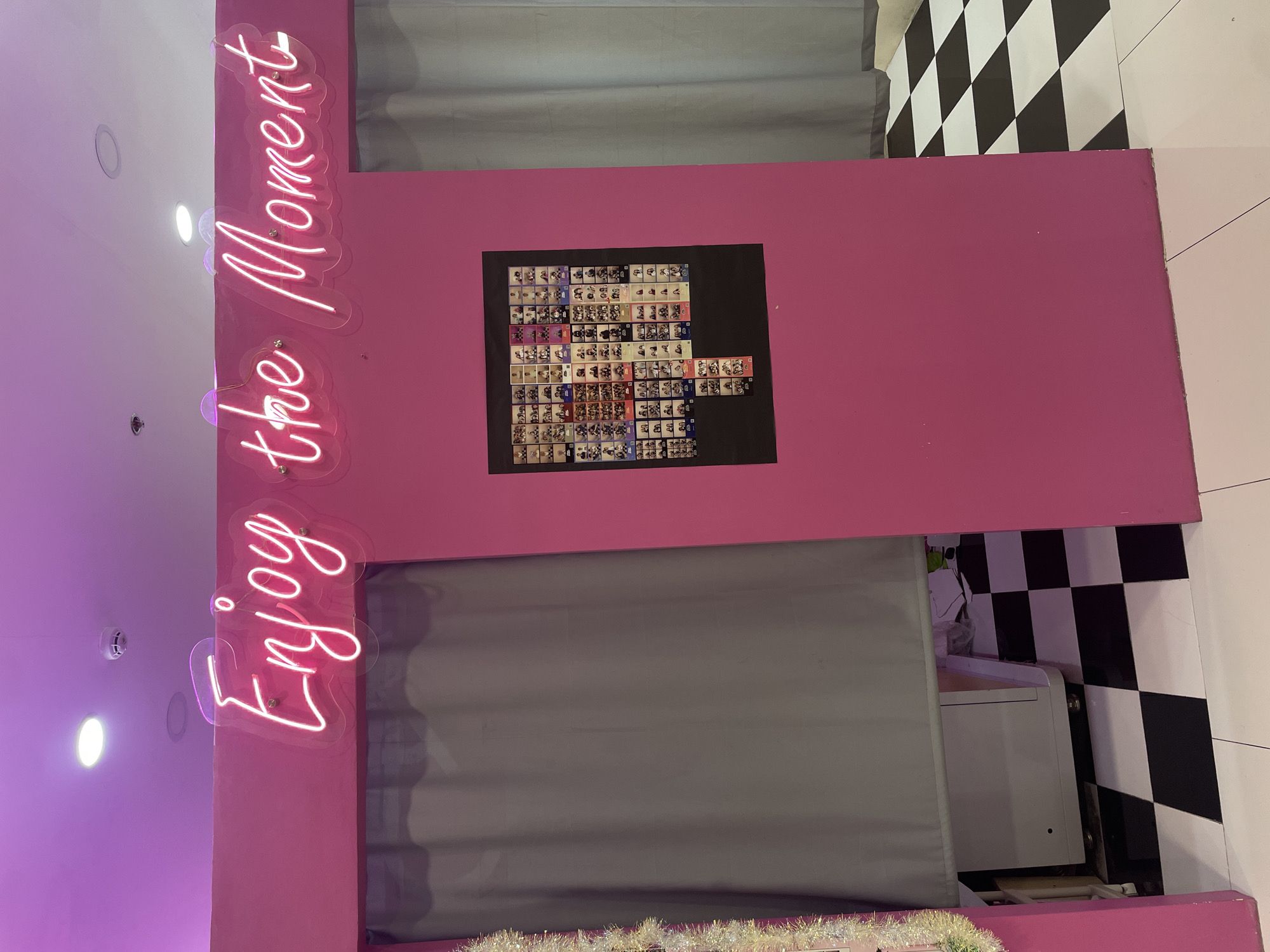 A neon sign reads "Enjoy the Moment" in pink neon lights against a bright pink wall in what appears to be a photo booth area, likely in Davao City, Philippines. To the left of the sign, a gray curtain hangs, suggesting a private space. On the pink wall, a collage of small photos is displayed, featuring individuals in various poses. Below the photo collage, a black checkered floor is visible. To the left of the photo booth, part of a silver photo booth machine and some festive tinsel are seen.