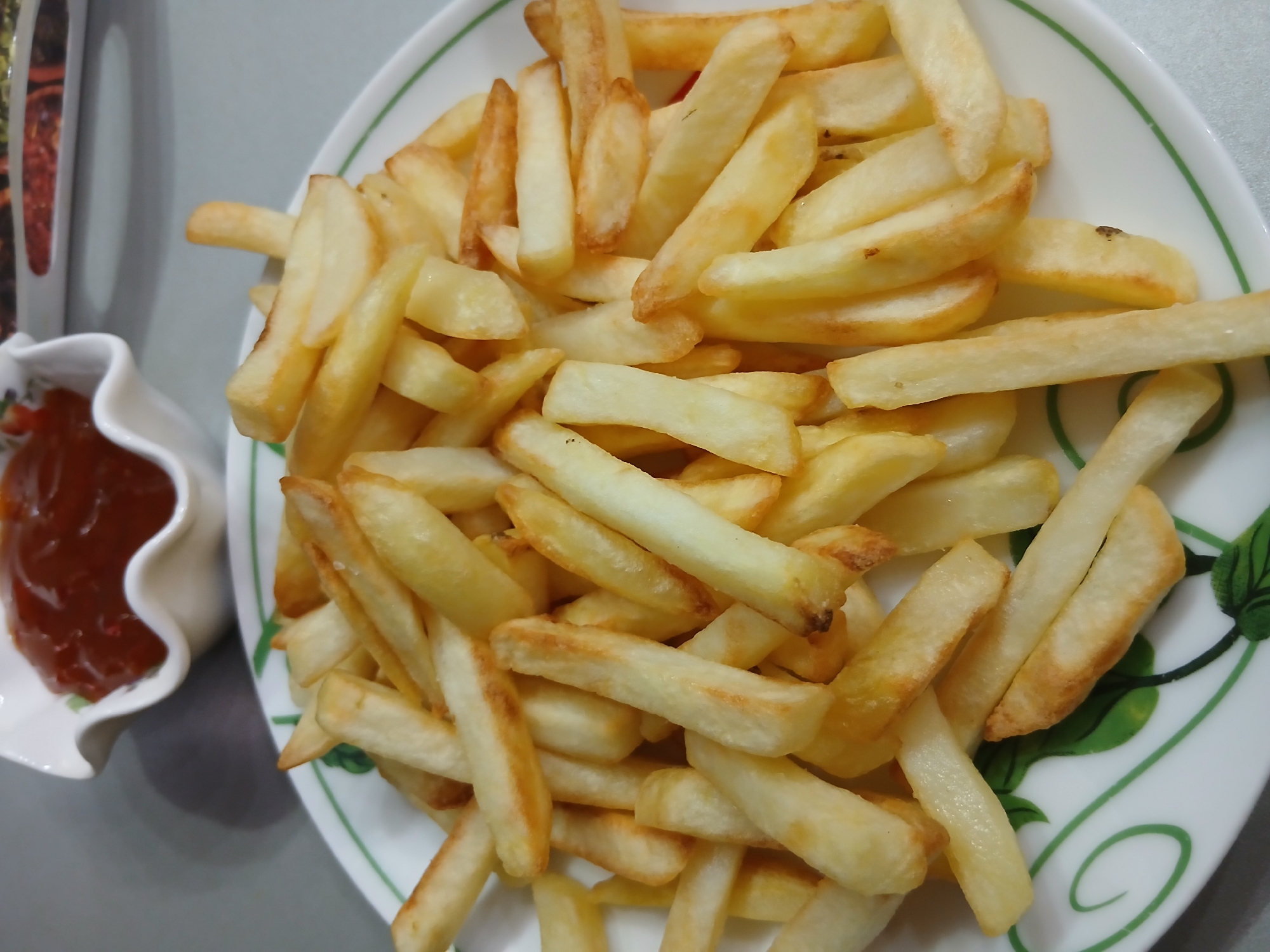 A plate of french fries is shown on a table, with a small bowl of ketchup in the background. The french fries are golden brown and appear to be freshly cooked. They are arranged in a pile on a white plate with a green decorative rim. The ketchup is a deep red color and is in a white, fluted bowl. The table appears to be a light gray surface.