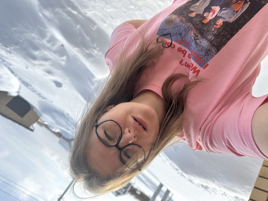 A female subject is depicted in a close-up, upward-angled selfie against a snow-covered outdoor background in Gornyi (Dvurechenskoe S/P), Russia. She has light brown hair, wears black-rimmed glasses, and a pink short-sleeved t-shirt featuring a graphic print with text partially visible as "Won't be cool I Ha be cool!" and an image of multiple figures. Her mouth is slightly open, and her eyes are partially closed. The background reveals a vast expanse of white snow with visible tracks and undulations, indicative of a winter environment. A light blue sky with minimal cloud cover is overhead. In the upper left, a grey metal structure, possibly a pole or part of a ski lift, is visible. In the upper right, a small, dark-roofed building is present on a distant snow-covered slope. The scene is brightly illuminated by natural light.