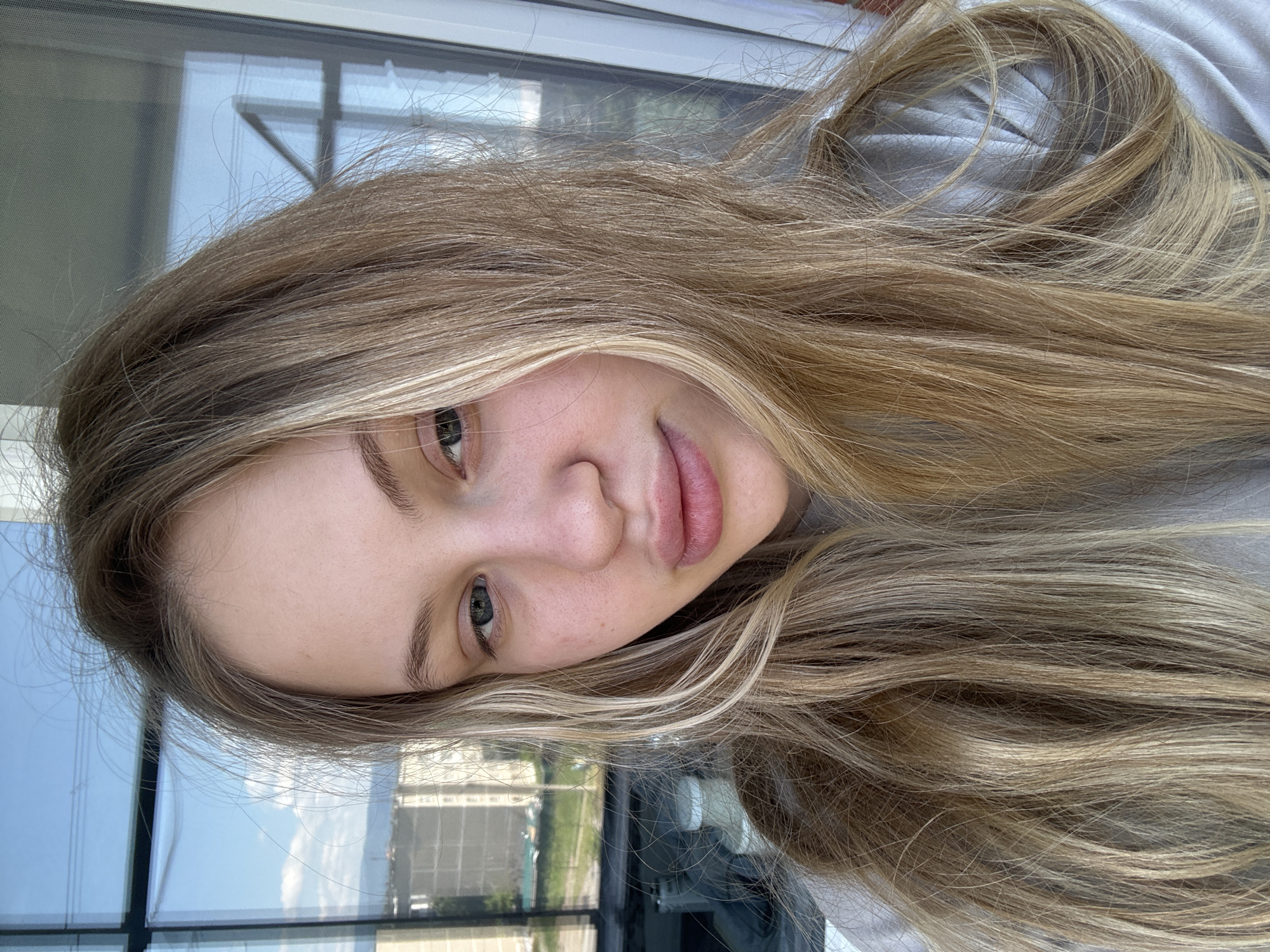 A medium shot shows a young Caucasian woman with long blonde hair and fair skin. She is looking towards the camera with a neutral expression. Her hair is light brown with blonde highlights, and it falls loosely around her face and shoulders. She is wearing a light-colored sweatshirt.
The background consists of a window with reflections of a cityscape and a blue sky with clouds. The architecture visible through the window appears to be apartment buildings, suggesting an urban environment. Based on the overall aesthetic and common building styles, this scene is likely located in Miass, Russia. The lighting is natural, coming from the window. There are no discernible objects or interactions in the foreground or midground, with the focus being solely on the woman's portrait.