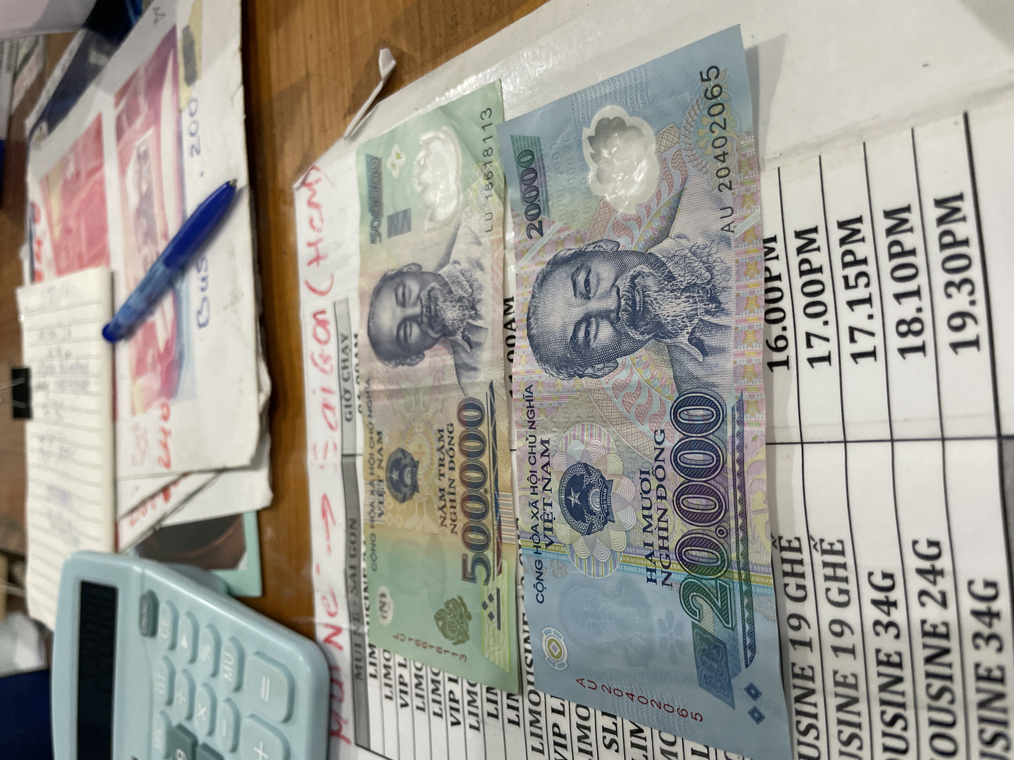 The image shows a desk with a light blue calculator on the left. In the center, there are two Vietnamese banknotes, a 500,000 Dong note and a 20,000 Dong note, laid out on a piece of paper with handwritten text. Behind the banknotes, there are other papers with writing and a blue pen. To the right, a stack of papers is visible. At the bottom of the image, a timetable with times and types of buses is partially visible, indicating a bus station or ticketing area. The overall scene suggests a transaction or administrative task taking place in what appears to be a busy environment.
The location is not specified in the image.