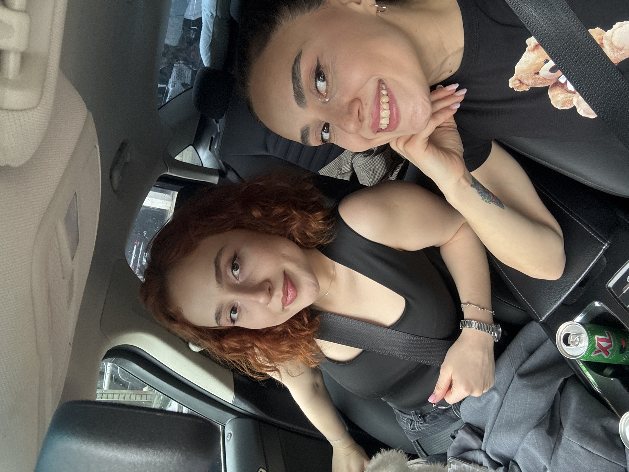 Two women are photographed inside a car. One woman has dark hair pulled back, wearing a black t-shirt with a teddy bear graphic, and has a tattoo on her arm. The other woman has red curly hair and is wearing a black tank top. Both women are wearing seatbelts. A soda can and gray clothing are visible on the car's center console. The location is identified as Tbilisi, Georgia based on visible buildings through the car window.